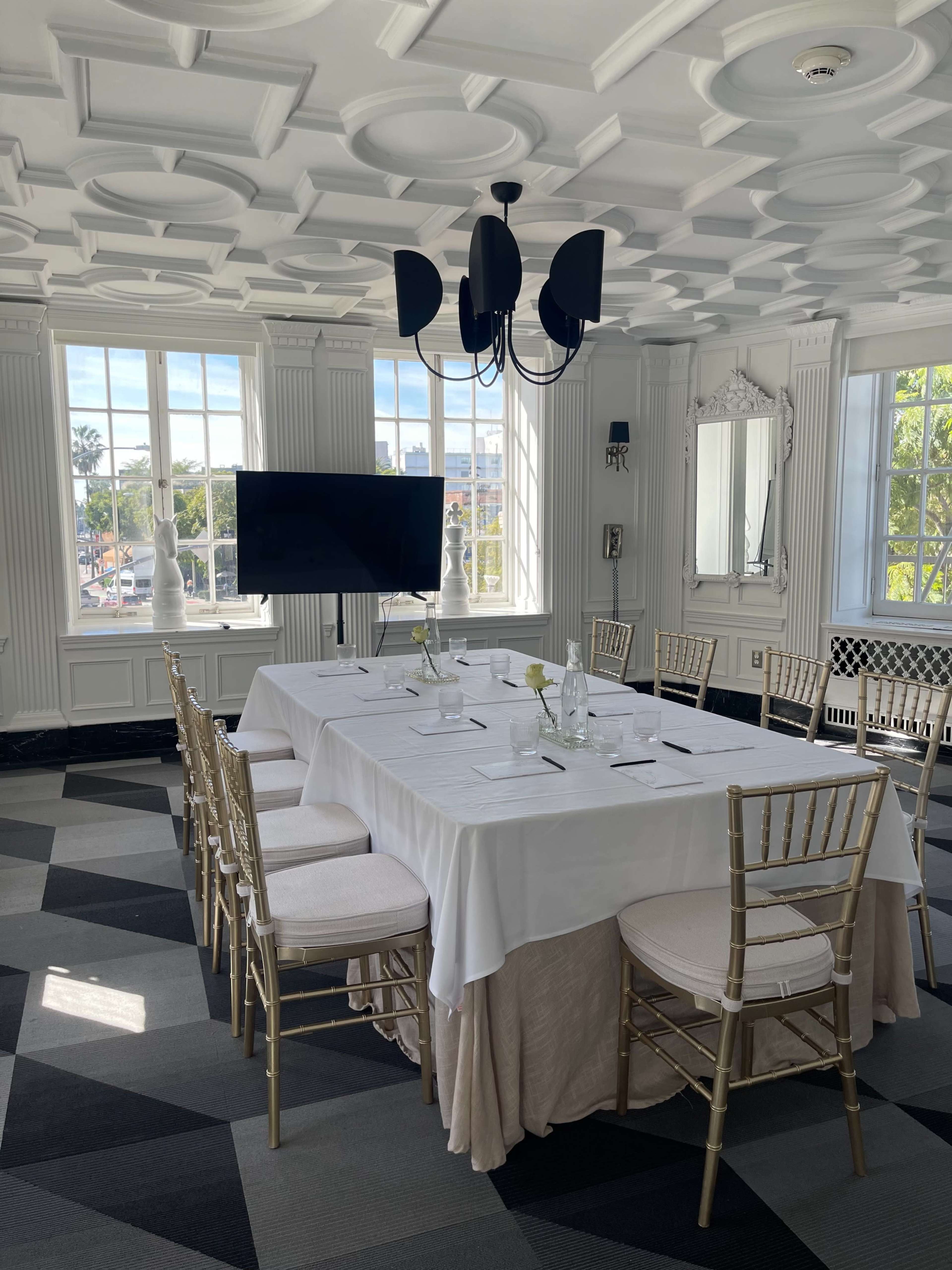 A bright meeting room features a large white table surrounded by gold chairs, with a television screen and large windows overlooking the city.