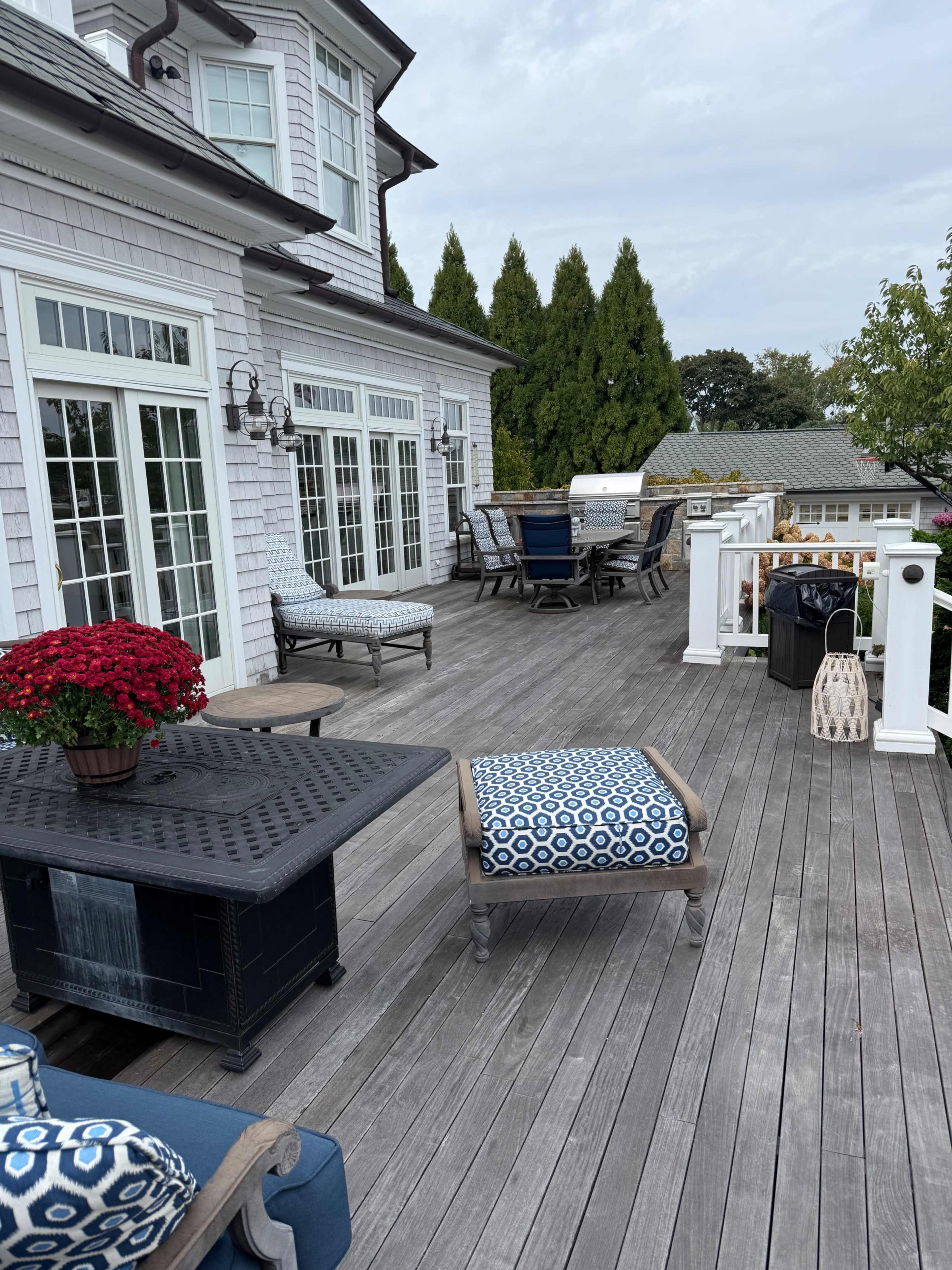 A spacious wooden deck features outdoor seating, a dining area, and potted flowers, adjacent to a house with large windows.