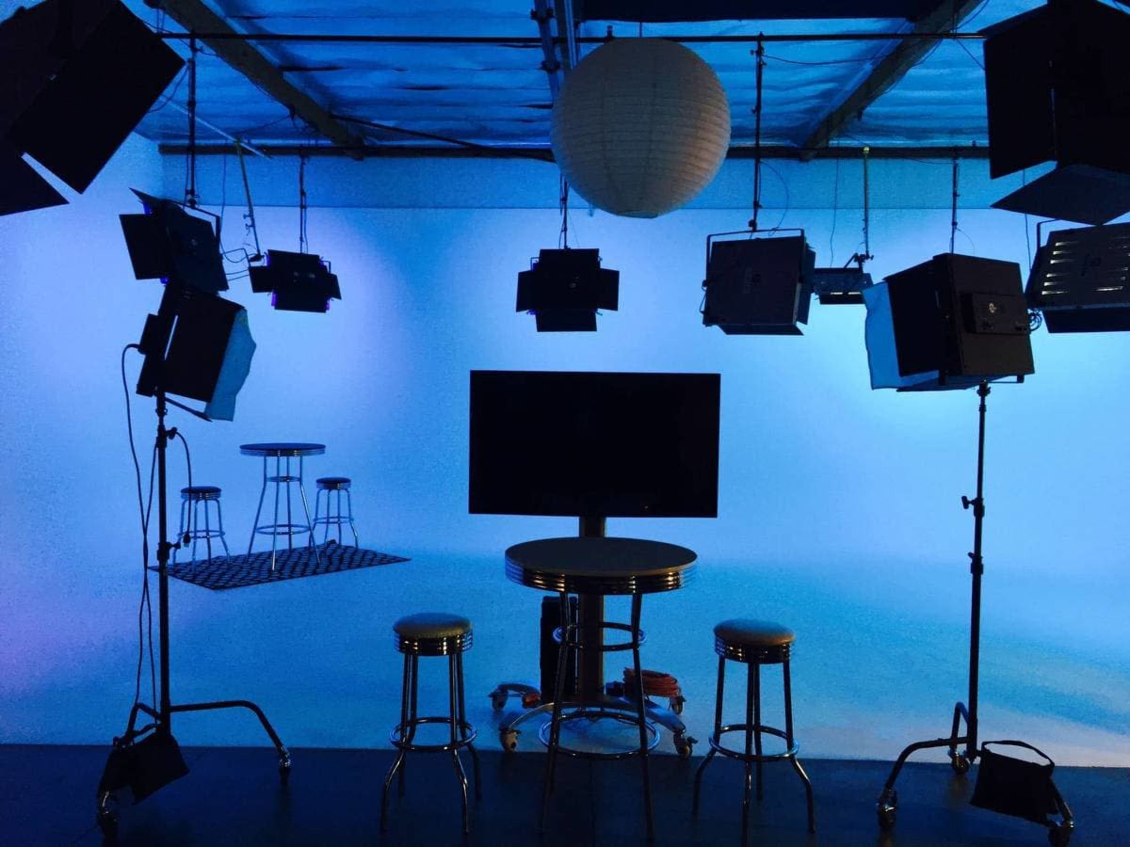 A video studio is set up with multiple lights, a large monitor on a table, and two stools against a blue backdrop.
