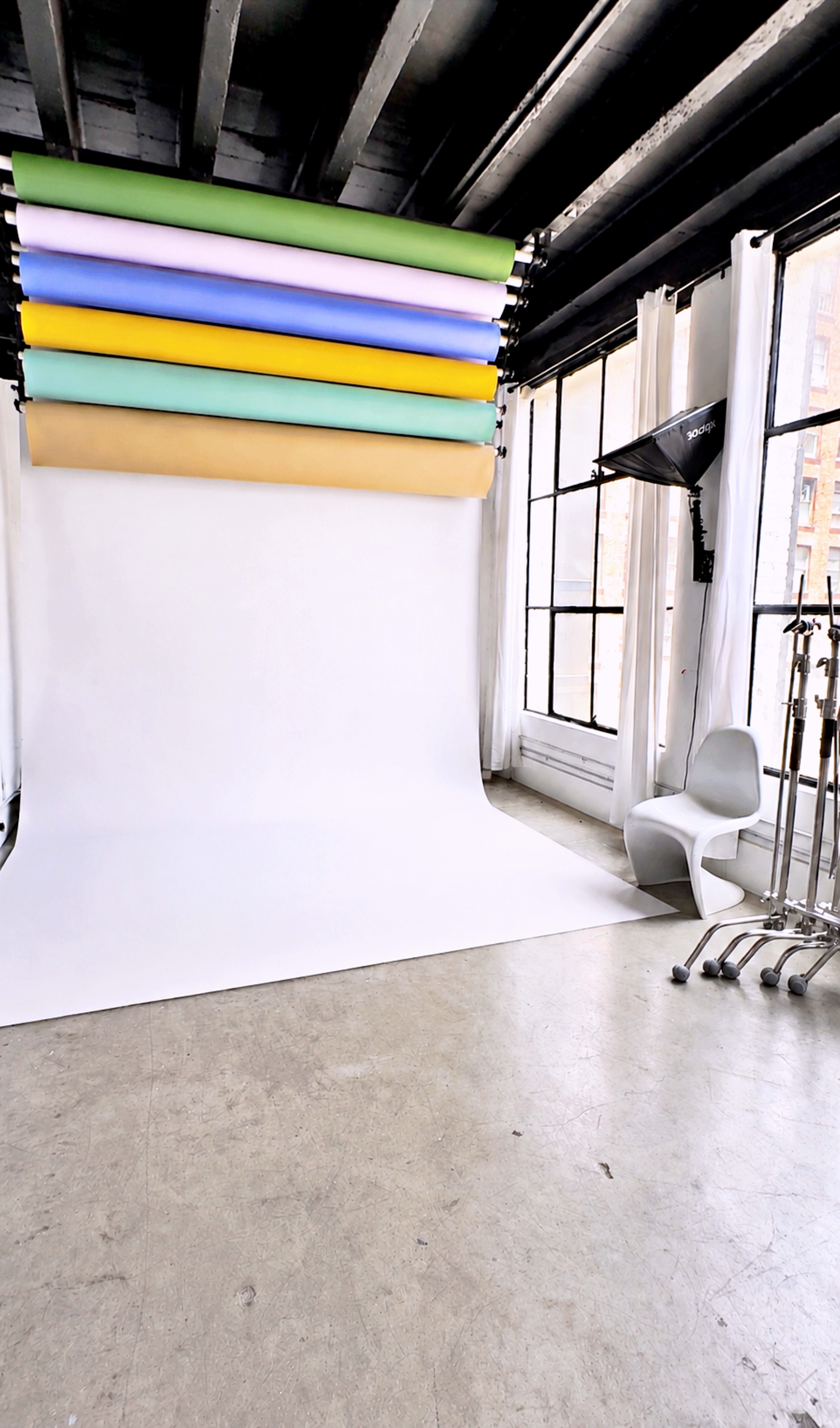 The image shows a photography studio with a white backdrop, several colored backdrop rolls overhead, and a chair positioned nearby.