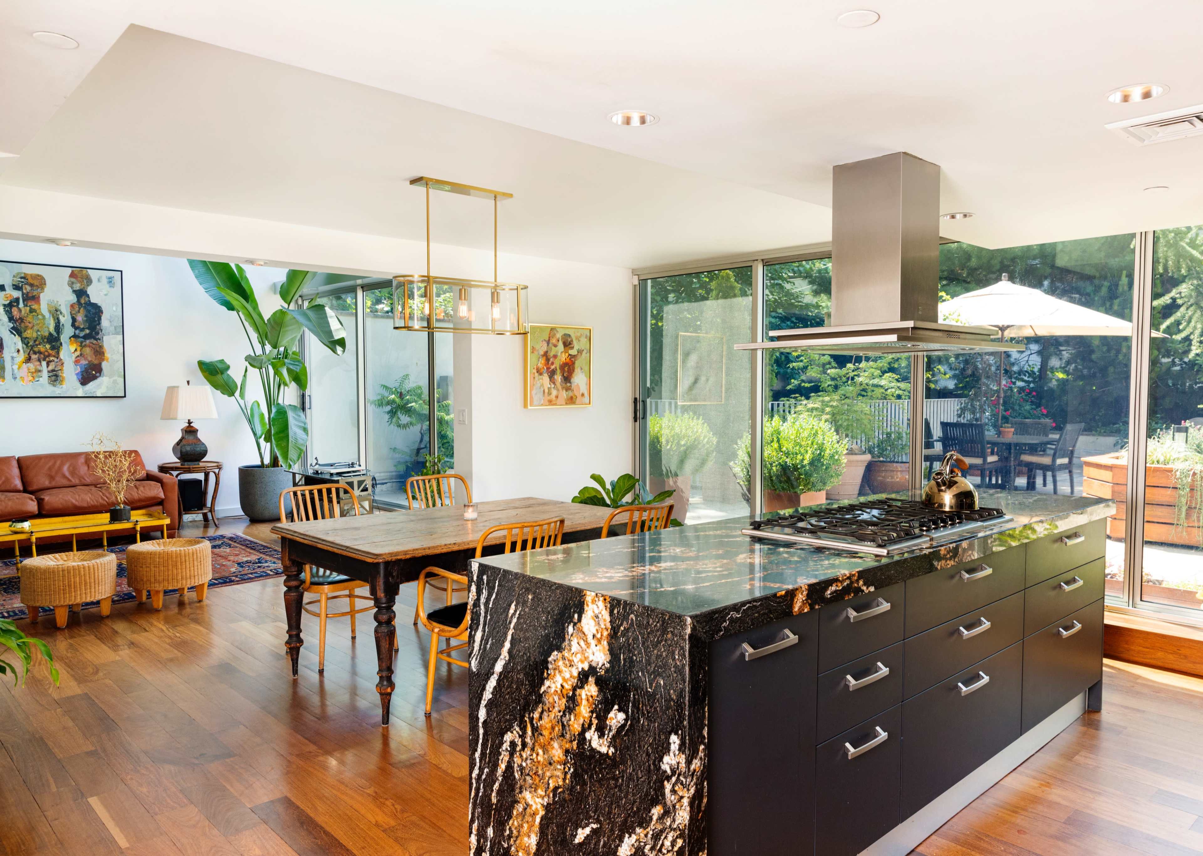 A modern kitchen featuring a large black and gold granite island, with wooden dining furniture and plants in a bright, open living space.