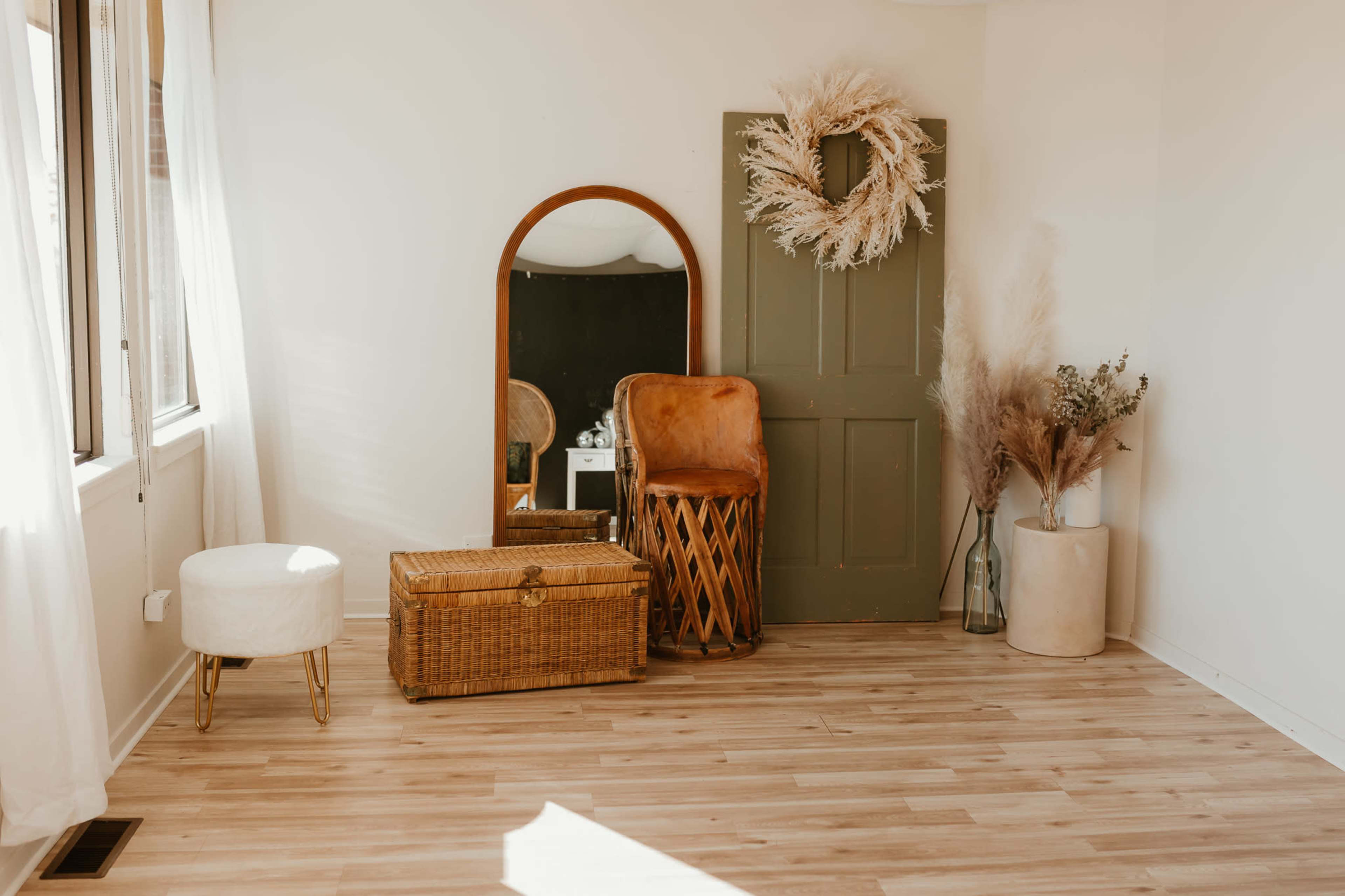 The scene features a minimalist room with a large mirror, a green door, a wicker chest, a tan chair, a small white stool, and decorative plants in vases.