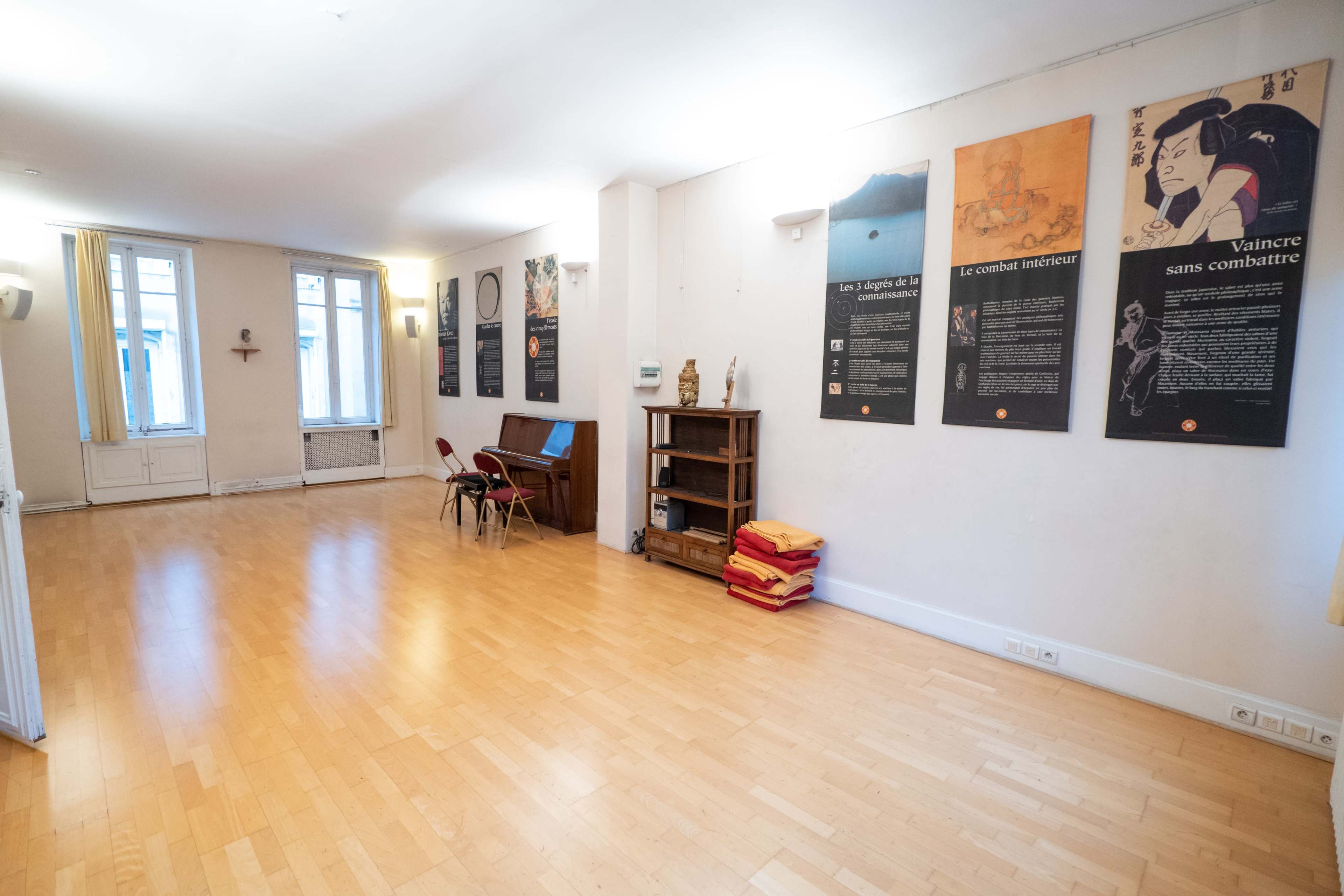 The image shows a spacious room with wooden flooring, featuring a desk, chairs, a bookshelf, and several informative posters on the walls.