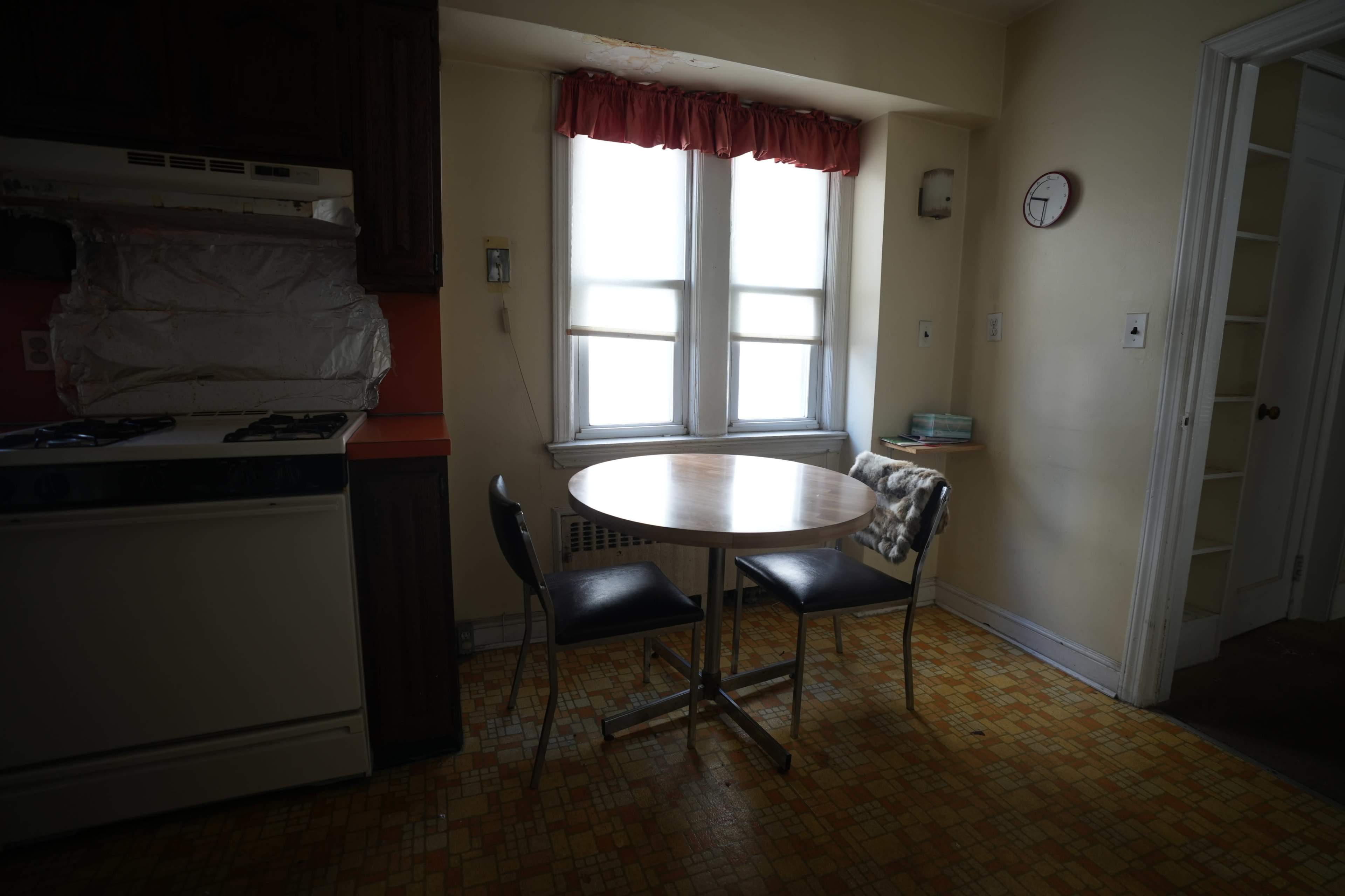 A small kitchen features a round table with two chairs, a stove, and a window adorned with red curtains.