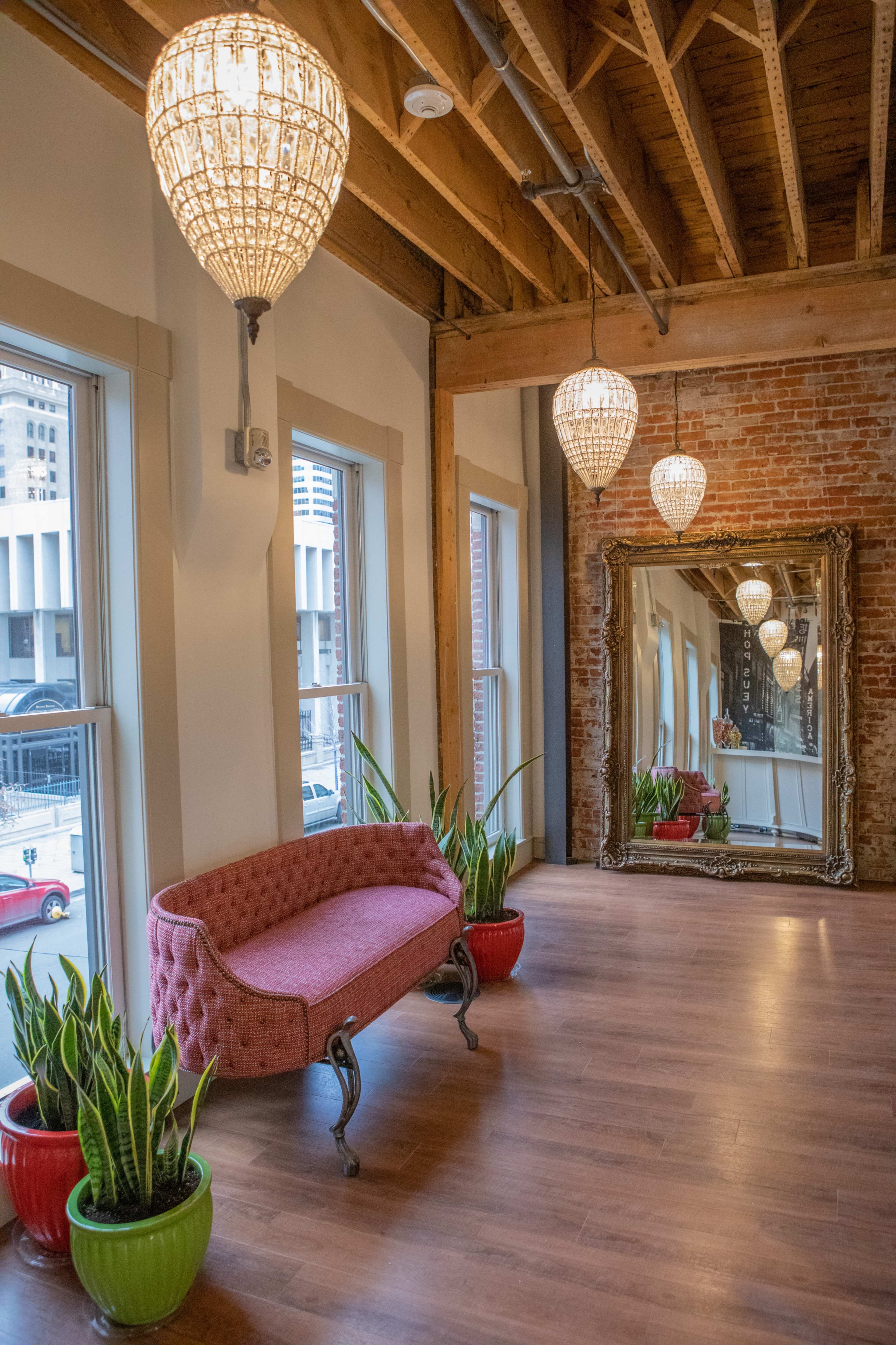 The room features a pink tufted sofa, decorative plants in red pots, a large mirror, and hanging chandeliers, all set against a backdrop of exposed brick walls and wooden beams.