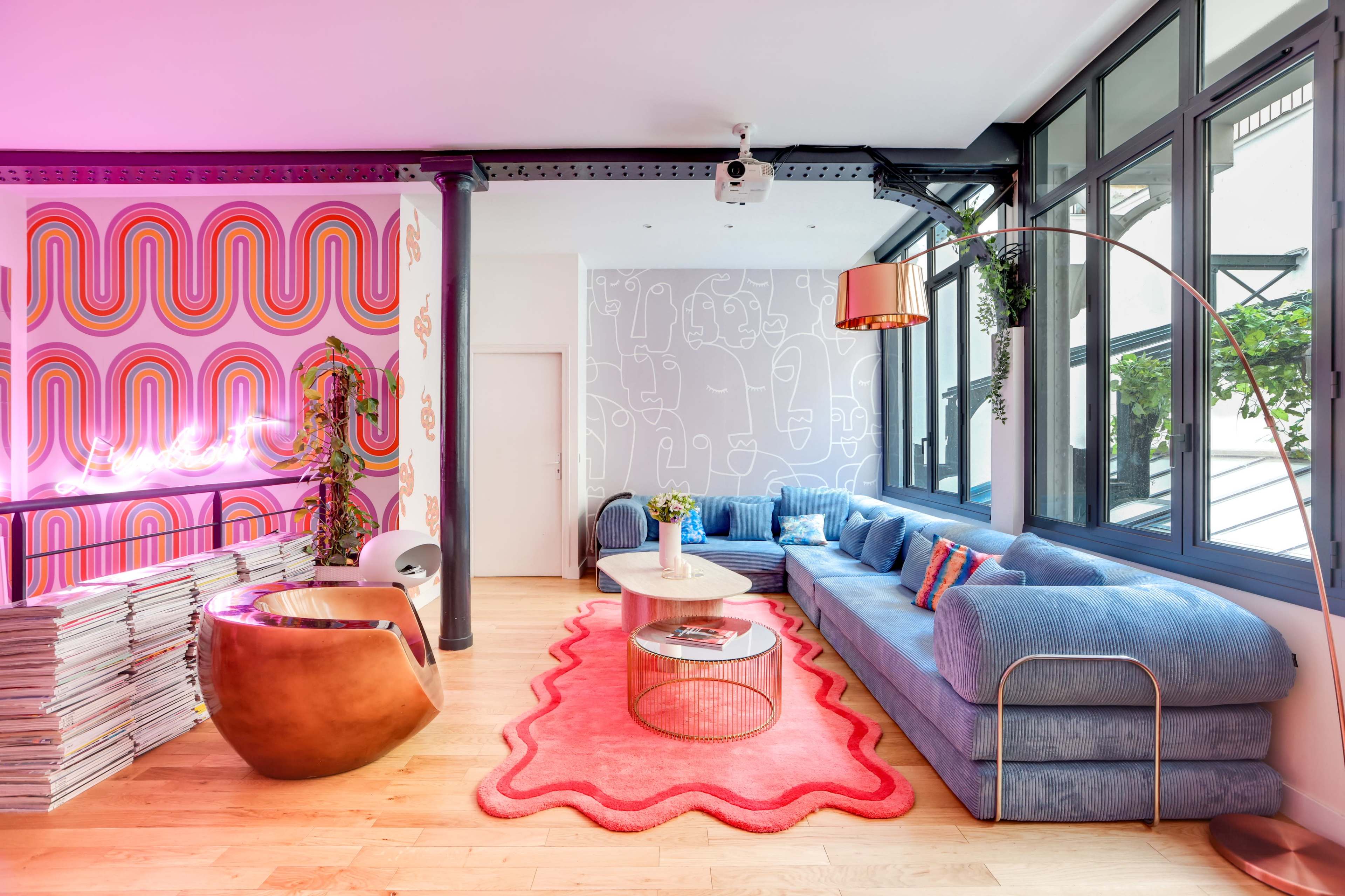 The image shows a modern living space featuring a blue sectional sofa, a circular coffee table, a brown curved chair, and colorful wall art, with large windows allowing natural light to enter.