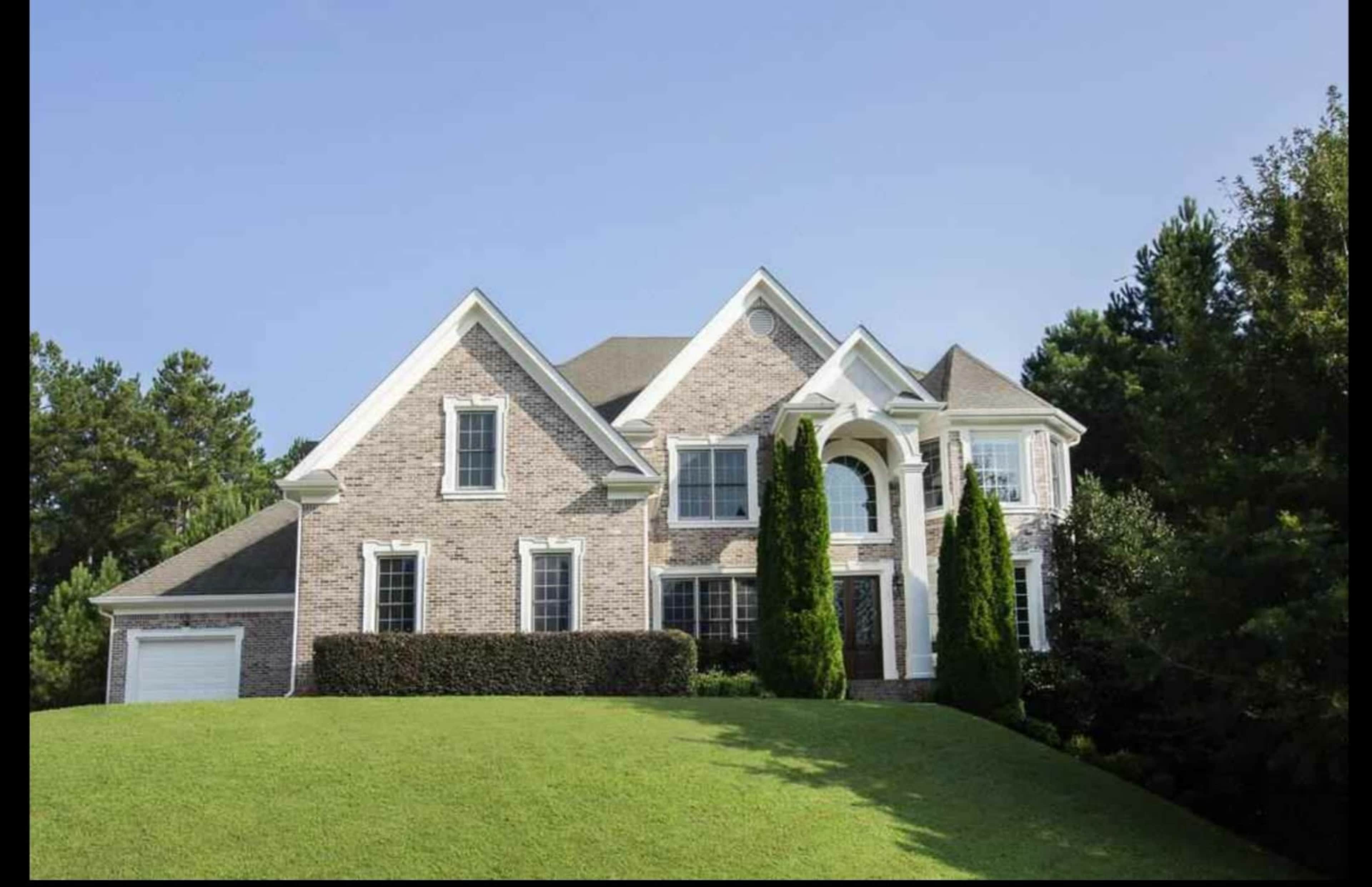 A large, two-story brick house with a symmetrical facade sits on a grassy hill surrounded by trees.