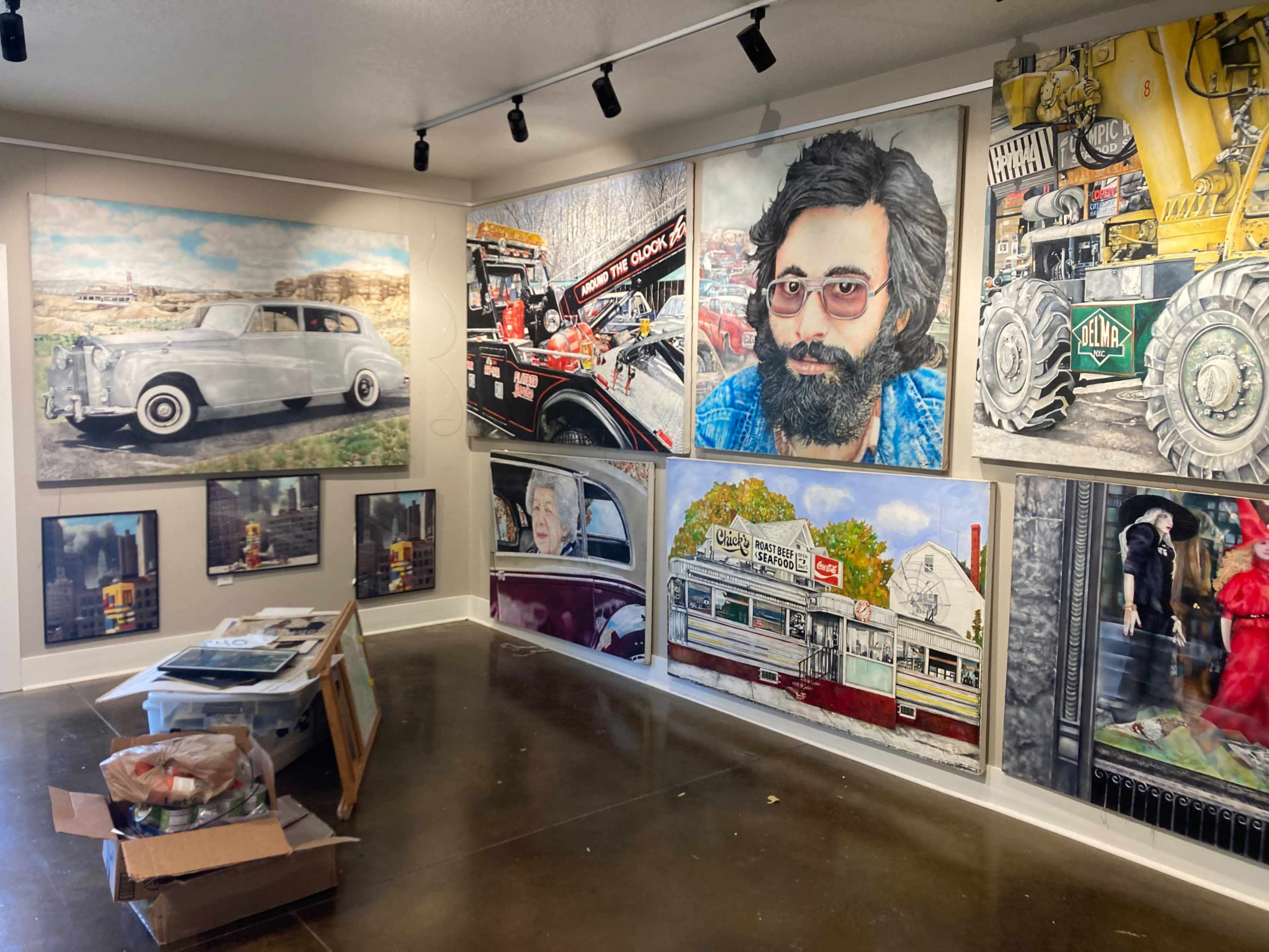 The image shows an art gallery featuring large portraits and various artworks displayed on the walls, with a table in the center holding materials and a cardboard box.