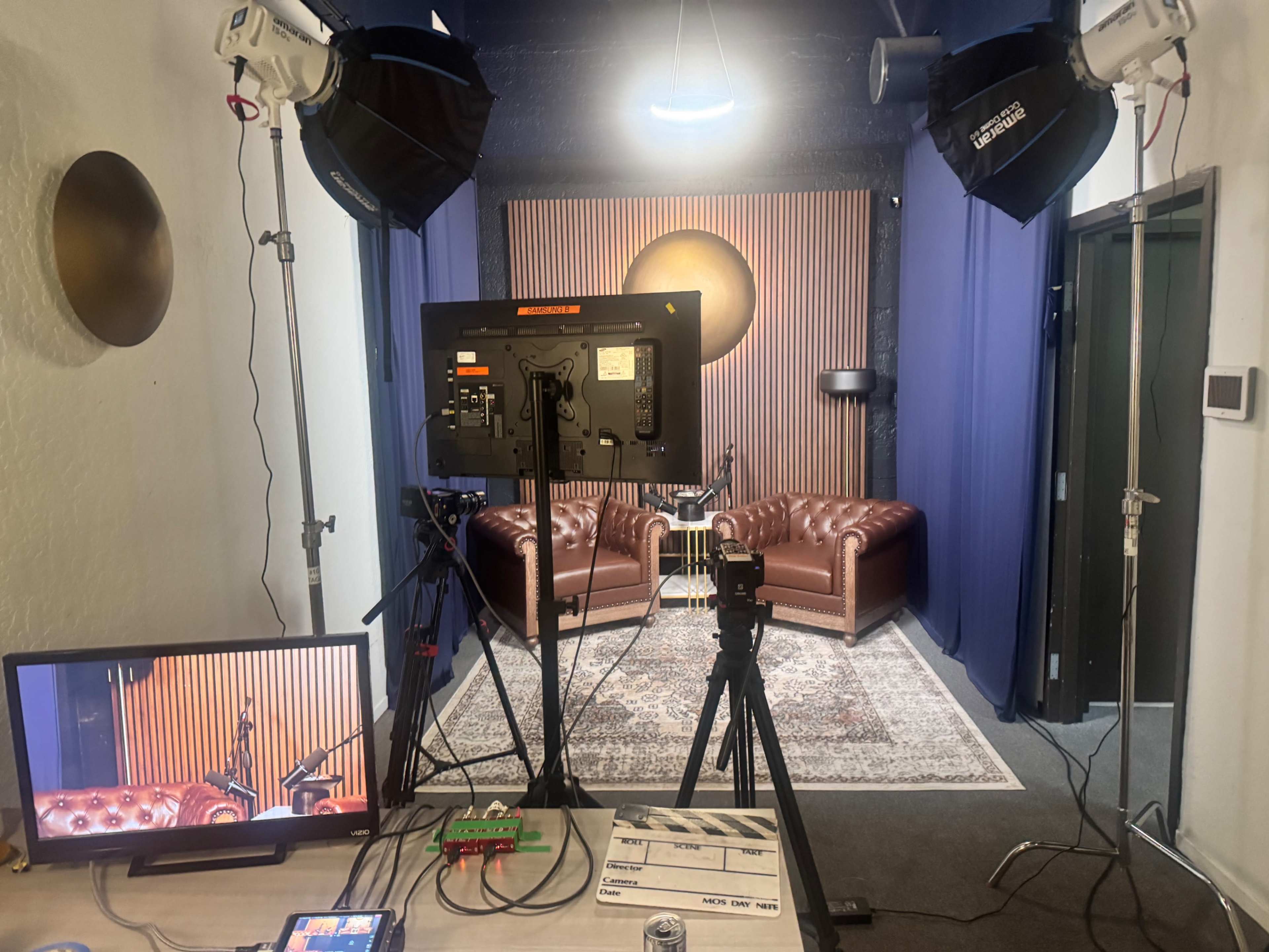 The image shows a video recording setup featuring two brown leather sofas against a backdrop of wooden slats, with studio lights and a camera gear positioned in front.