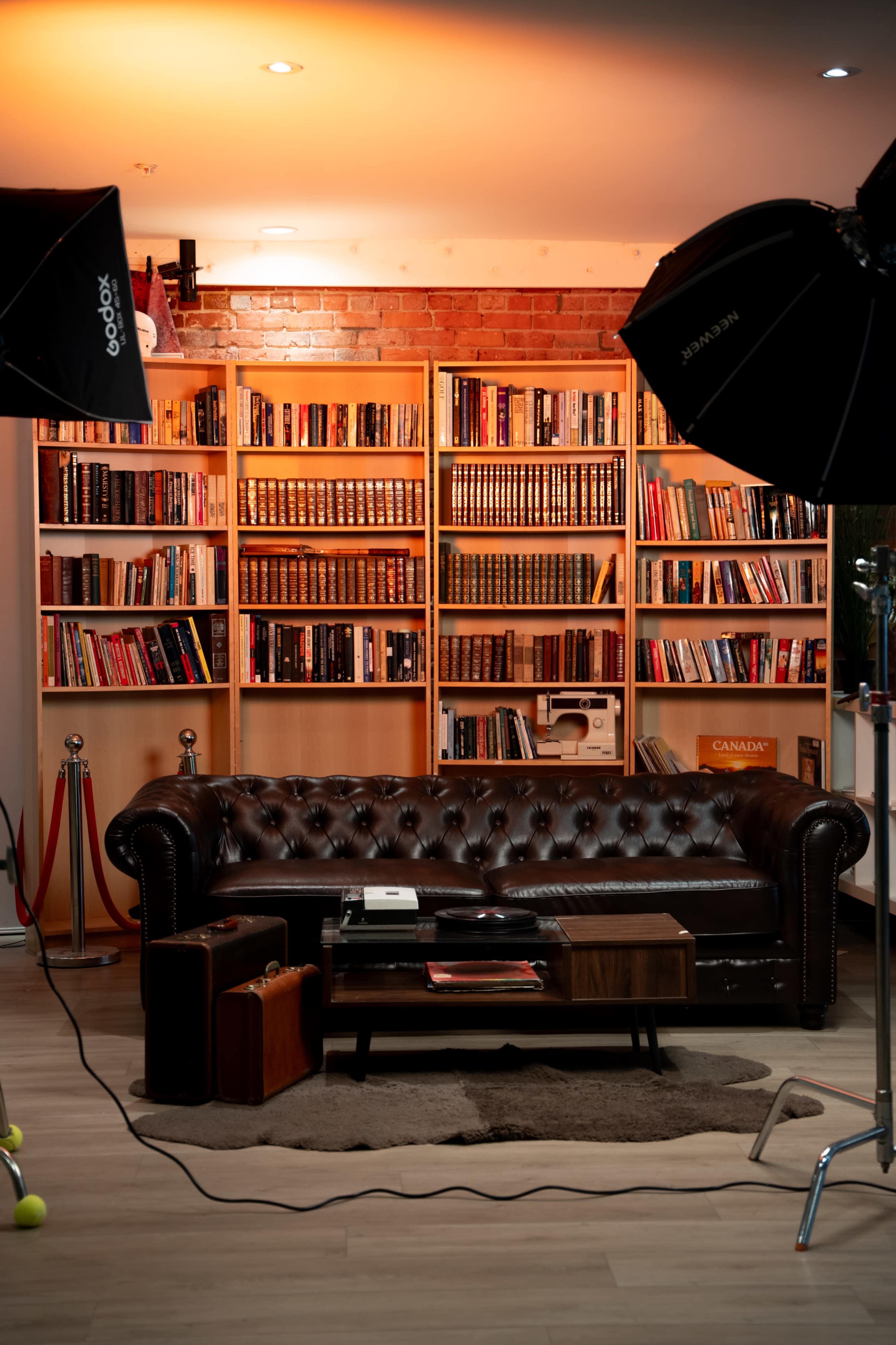 The image features a brown leather chesterfield sofa facing a wooden coffee table, with a bookshelf filled with books in the background and studio lights positioned above.