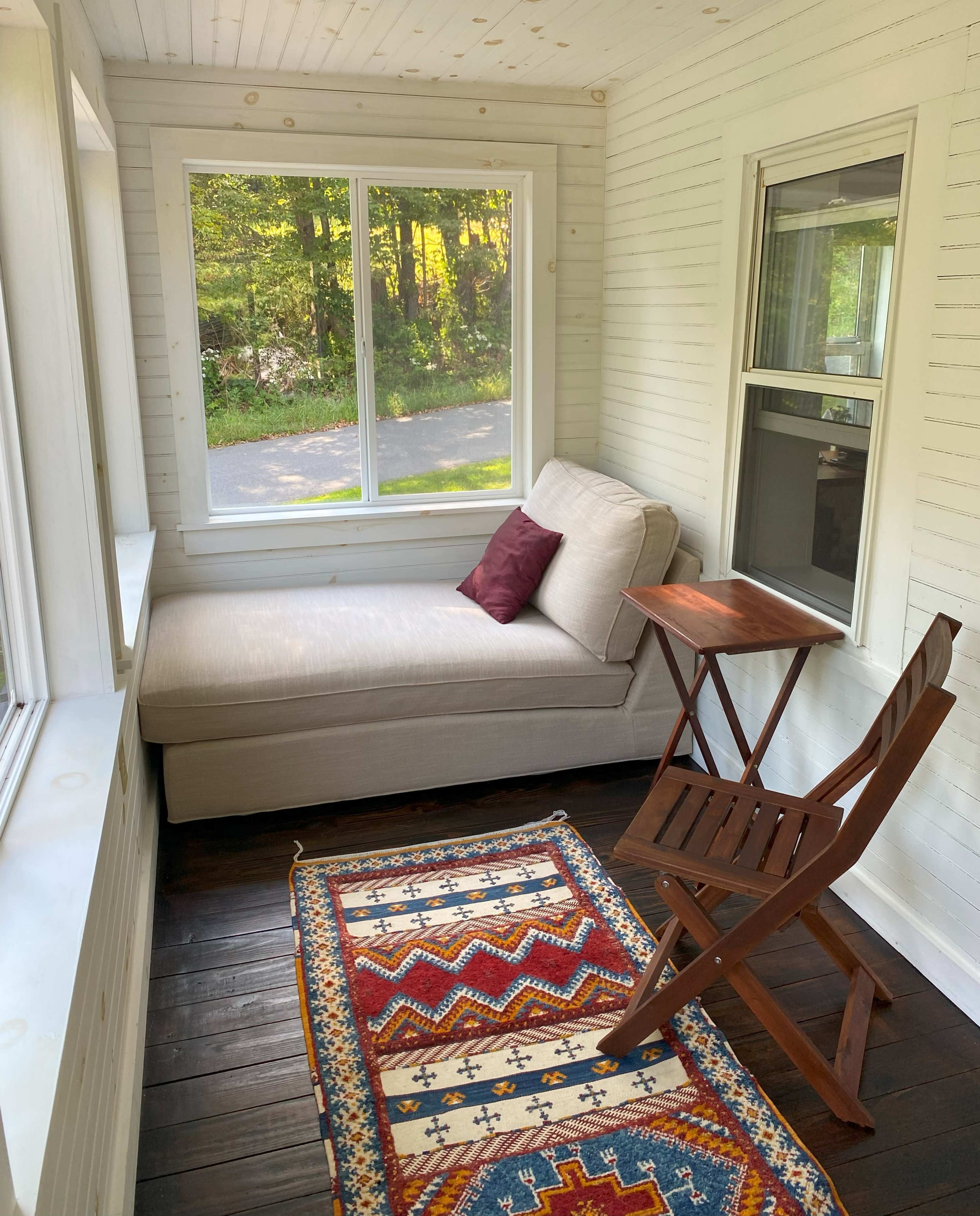 A sunlit room features a beige chaise lounge, a small wooden folding table, and a colorful area rug on a dark wooden floor, with windows offering a view of greenery outside.