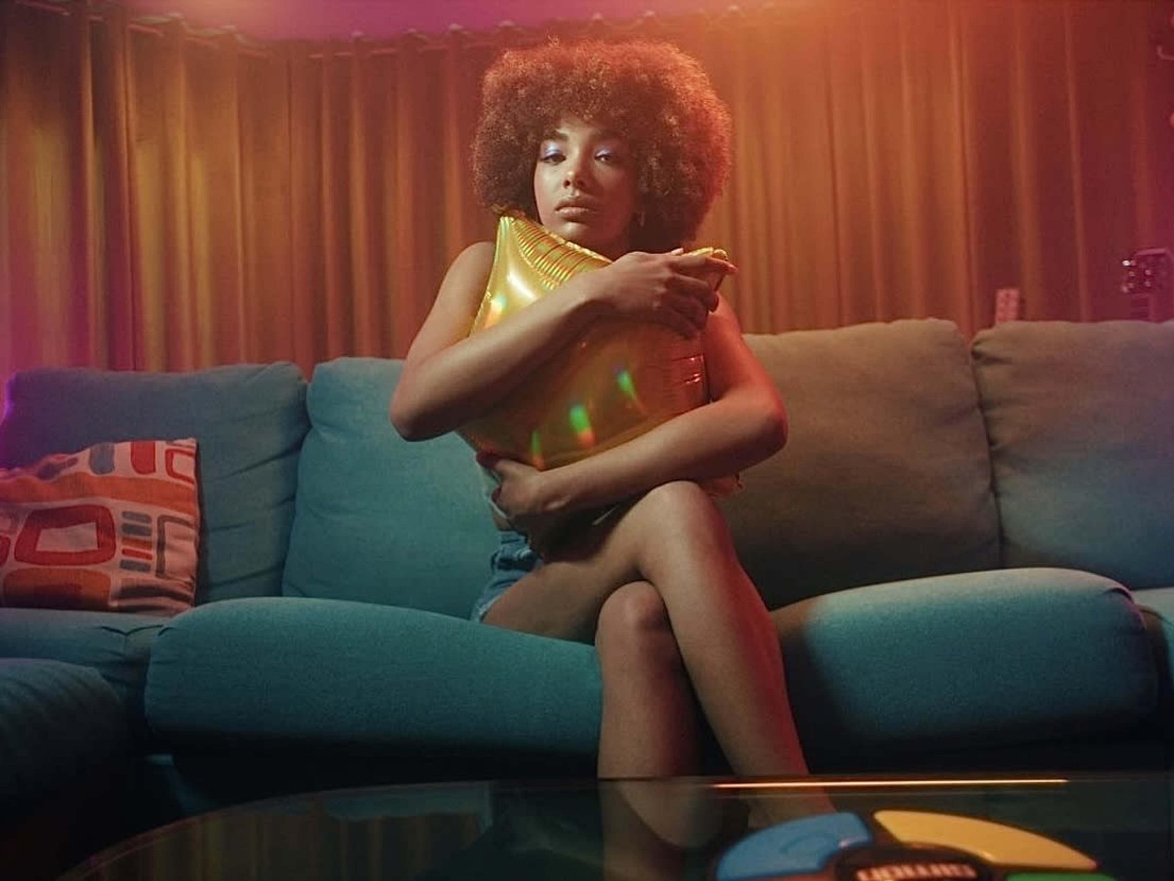 A person with curly hair sits on a couch, hugging a gold pillow while surrounded by colorful lighting.
