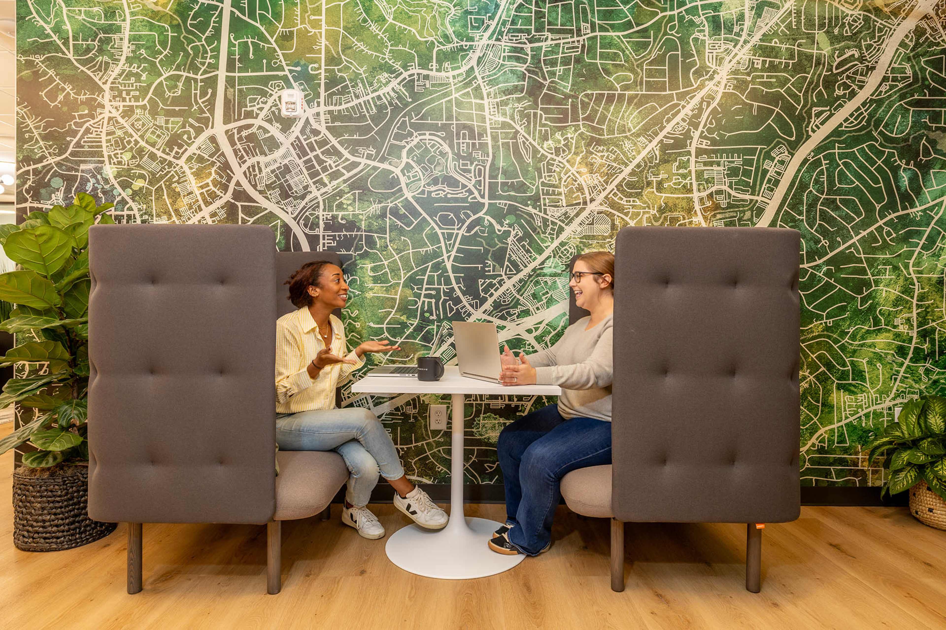 Two women are engaged in conversation at a small table in a cafe-like setting, surrounded by a large map-themed wall and indoor plants.
