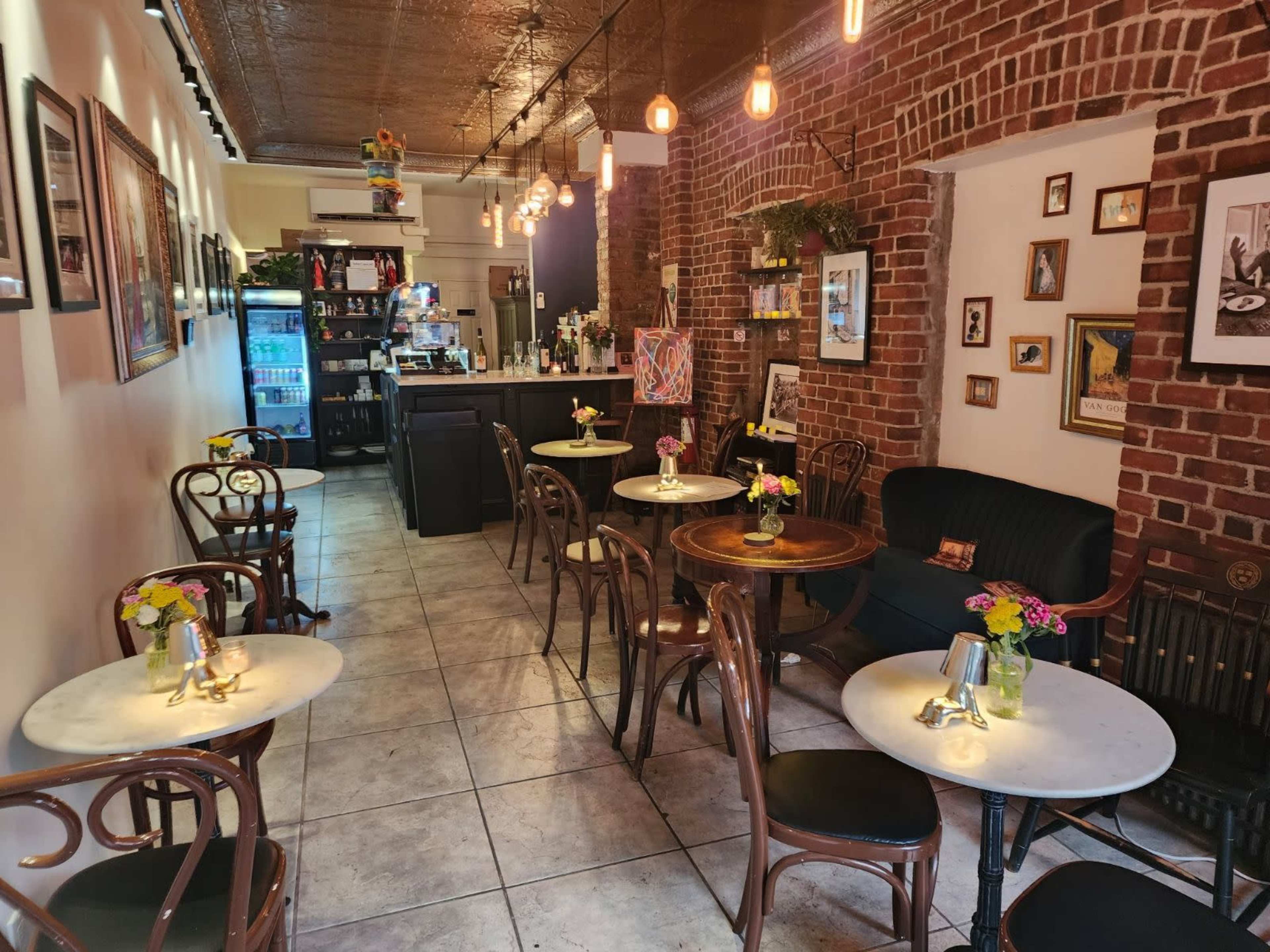The image shows a cozy café interior with brick walls, small tables adorned with flower vases, and a counter in the background.