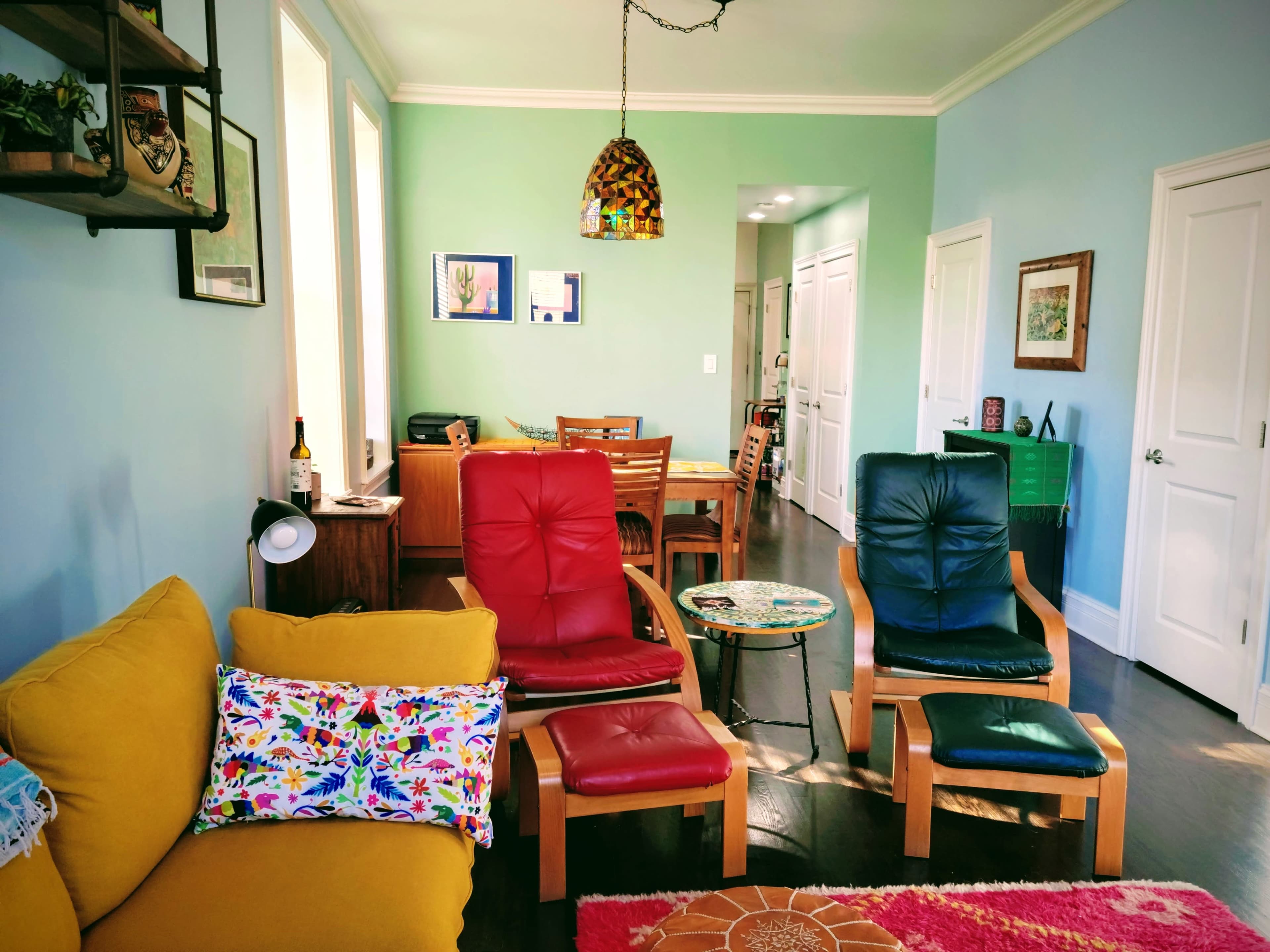 A living room setting with yellow, red, and blue chairs, a small round table, and a dining area visible in the background.