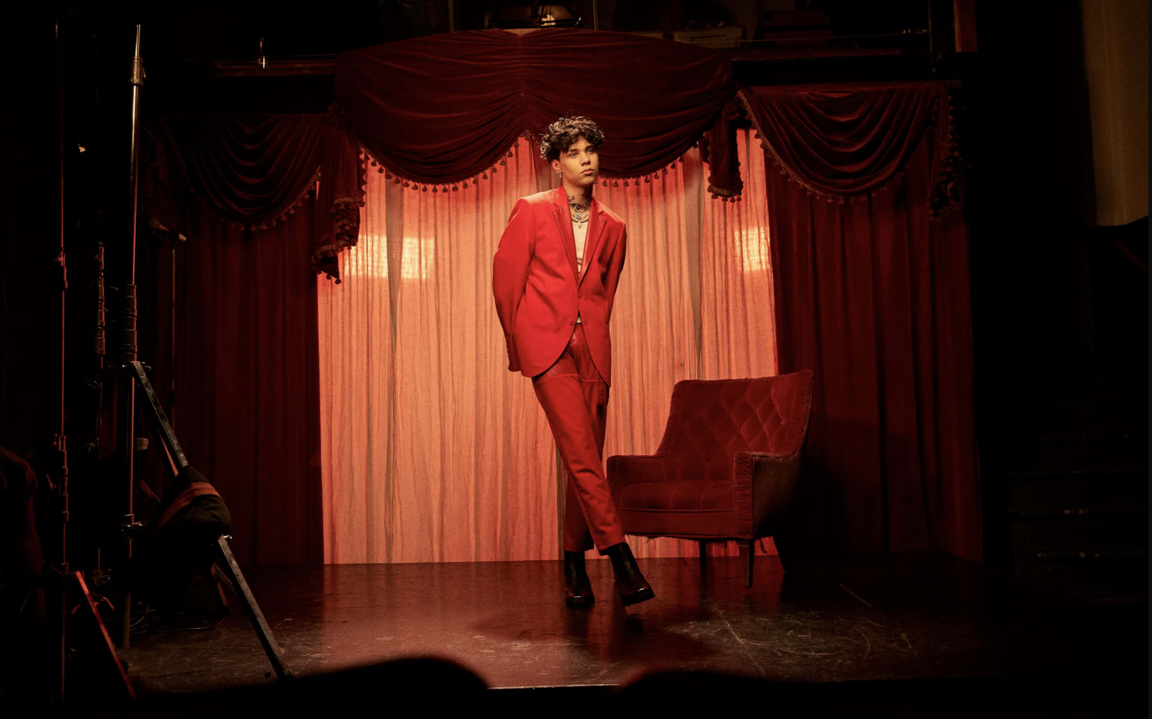 A person in a red suit stands confidently on a stage in front of a red curtain, next to an empty red chair.