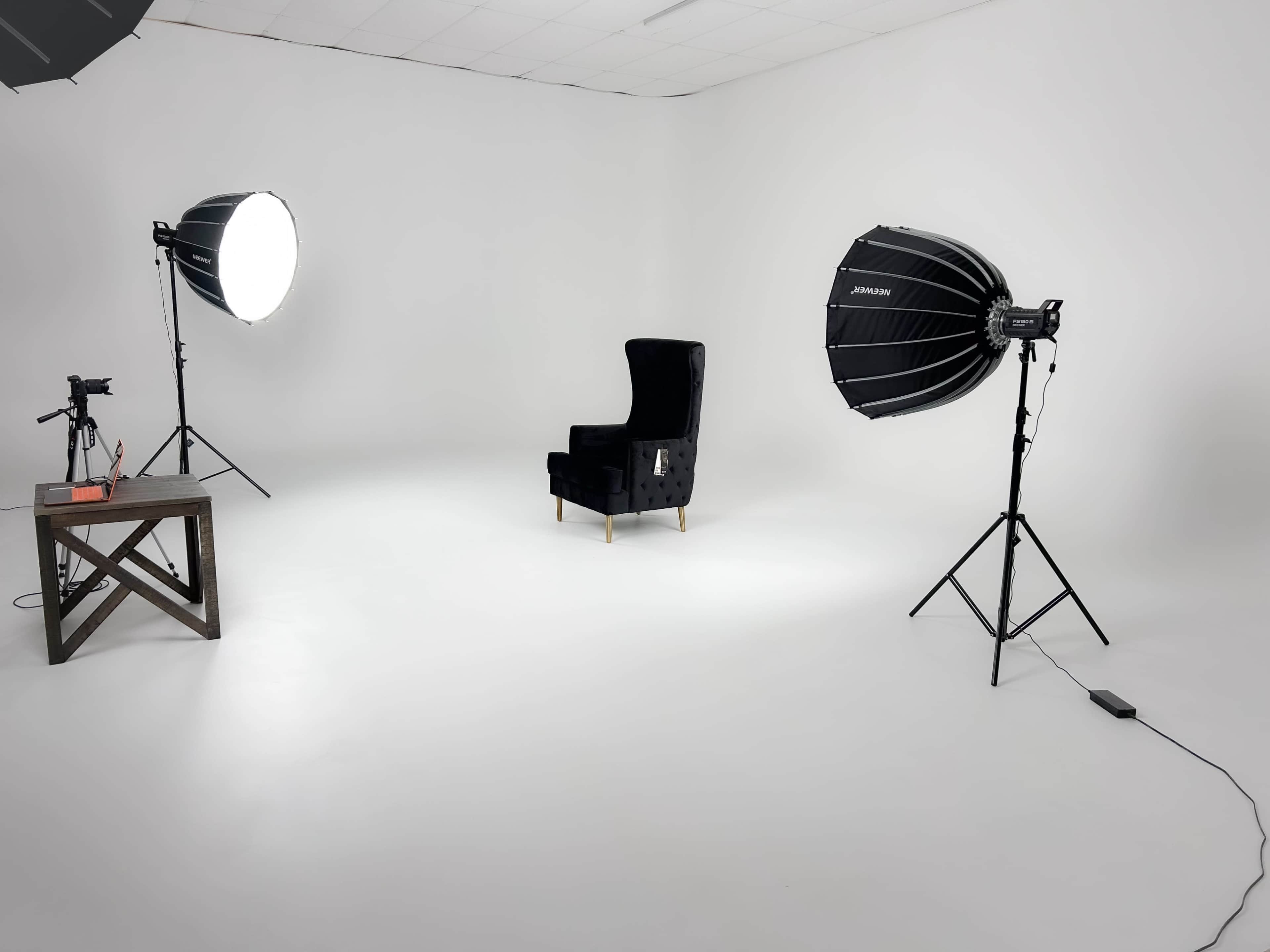 A minimalist studio setup featuring a single black armchair, two large softbox lights, and a camera on a table against a white backdrop.