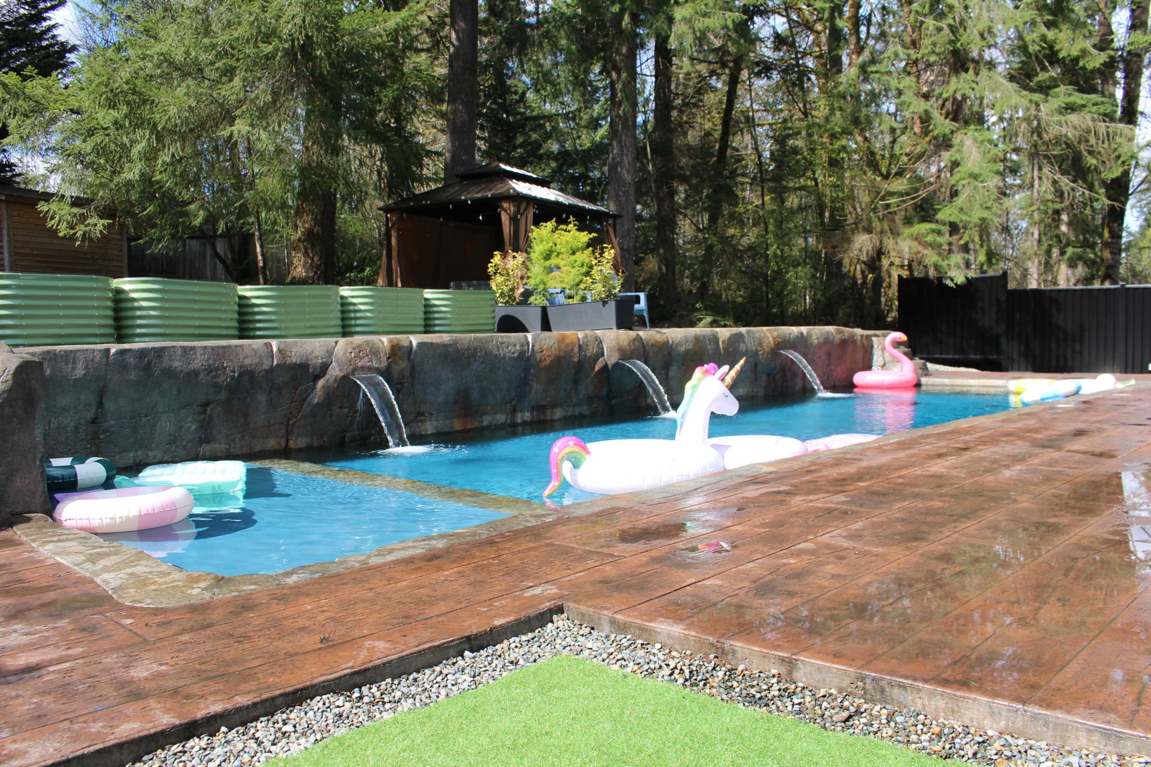 The image shows a swimming pool surrounded by wooden decking, featuring inflatable unicorns and flamingos, with waterfalls cascading from a rock formation and greenery in the background.
