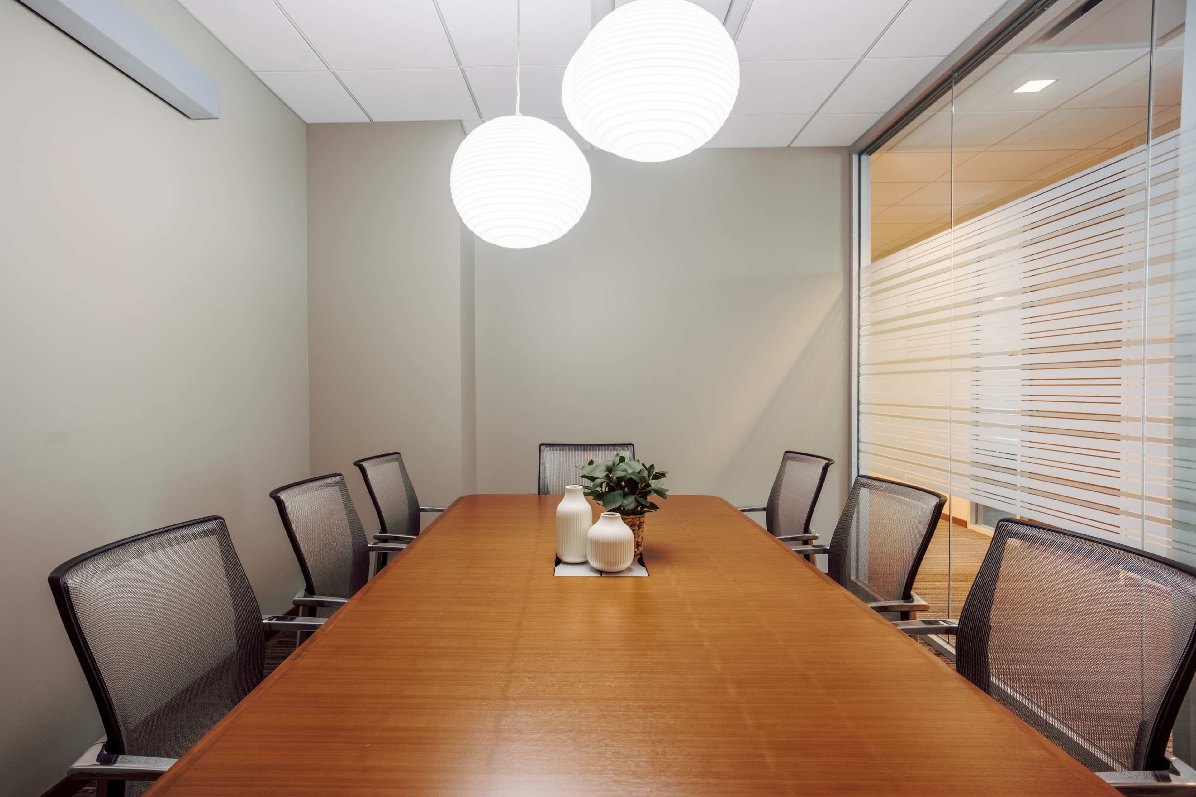 The image shows a modern conference room with a long wooden table surrounded by six chairs and two pendant lights hanging from the ceiling.