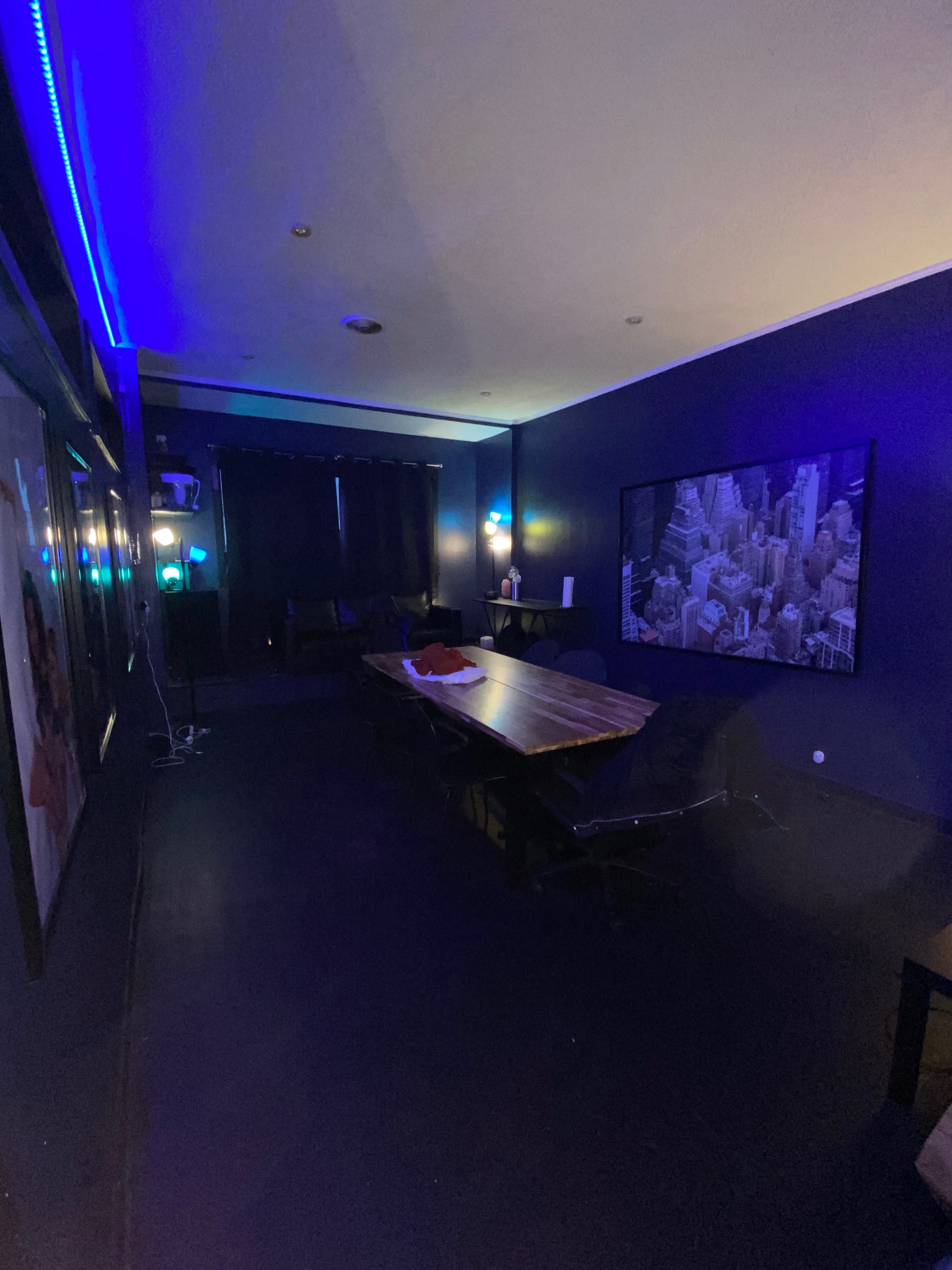 A dimly lit room with a large table, dark seating, and a wall-mounted screen displaying a cityscape.