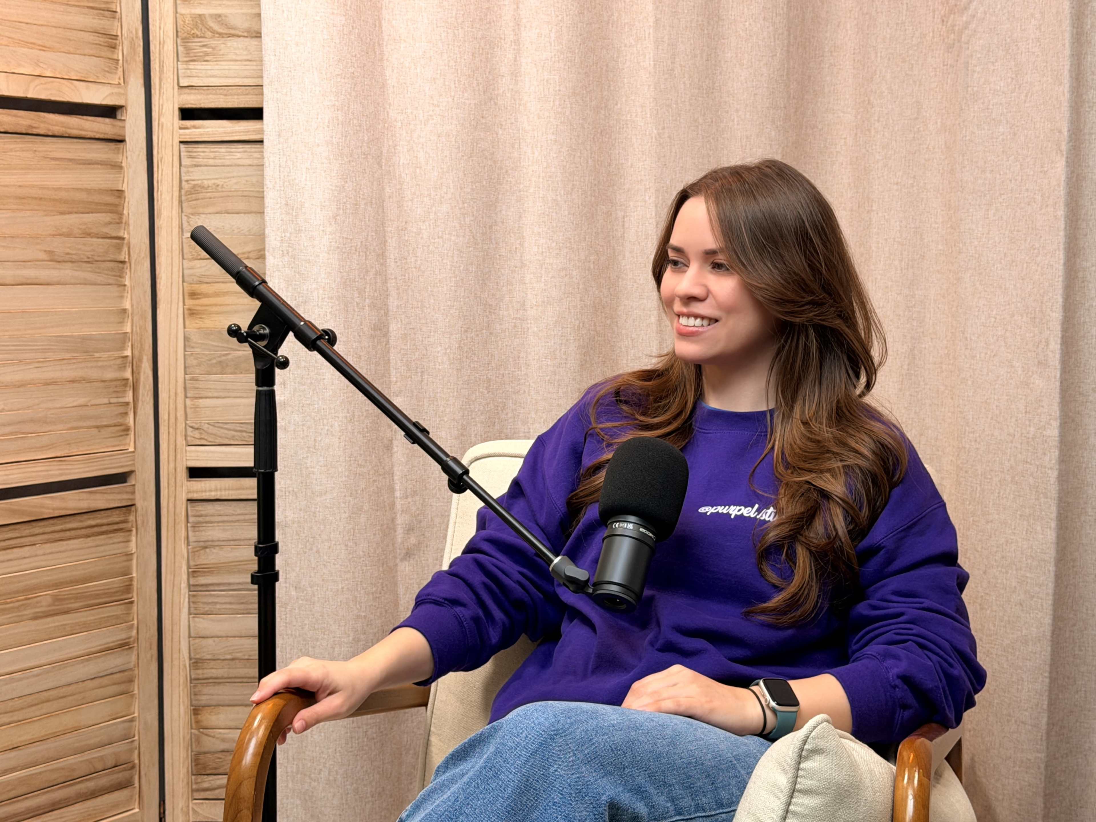 A woman with long hair sits in a chair, smiling while wearing a purple sweatshirt and a microphone is positioned in front of her.