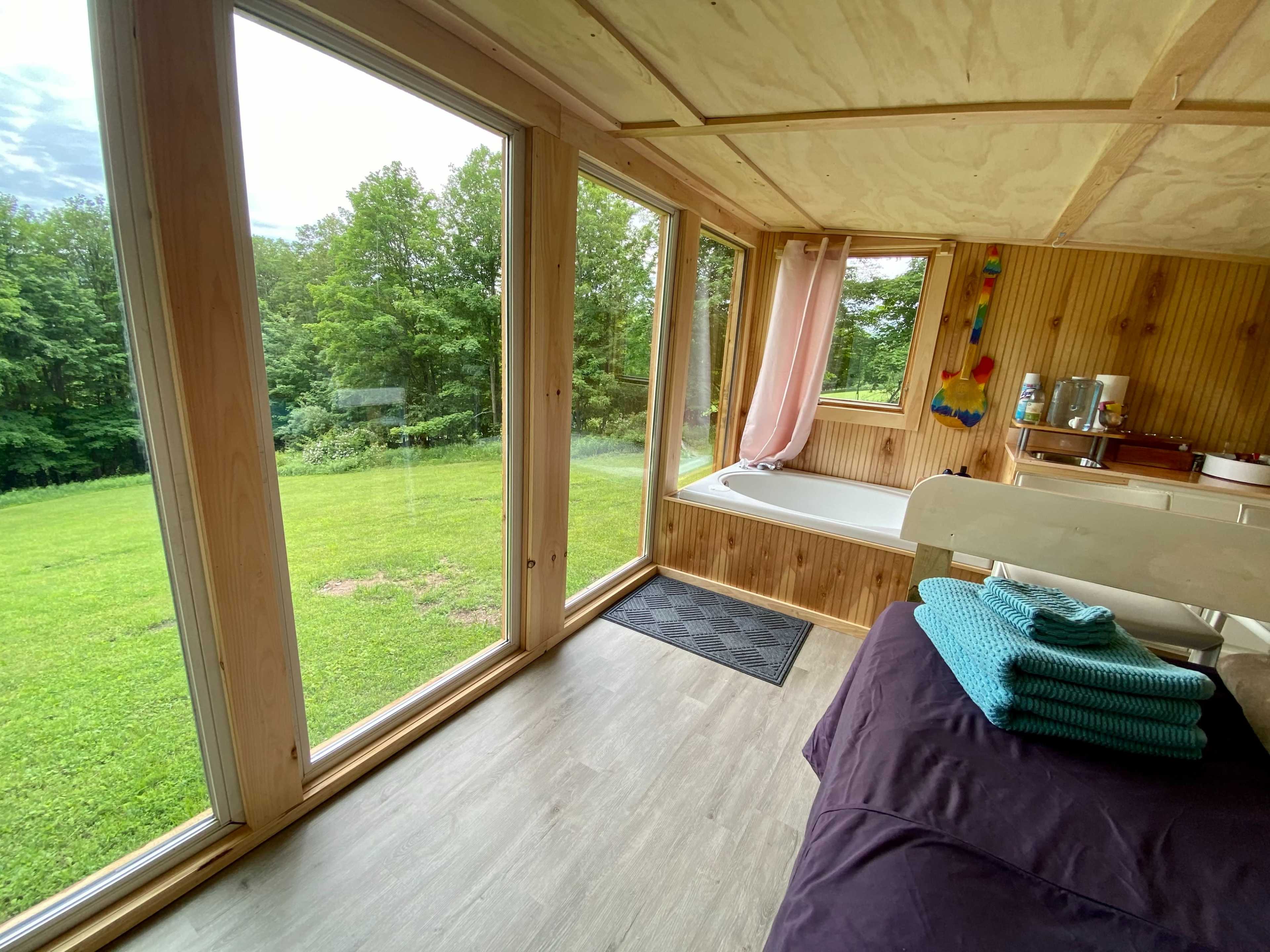 A bright room with large windows overlooks a green lawn and features a bed, a bathtub, and neatly stacked towels.