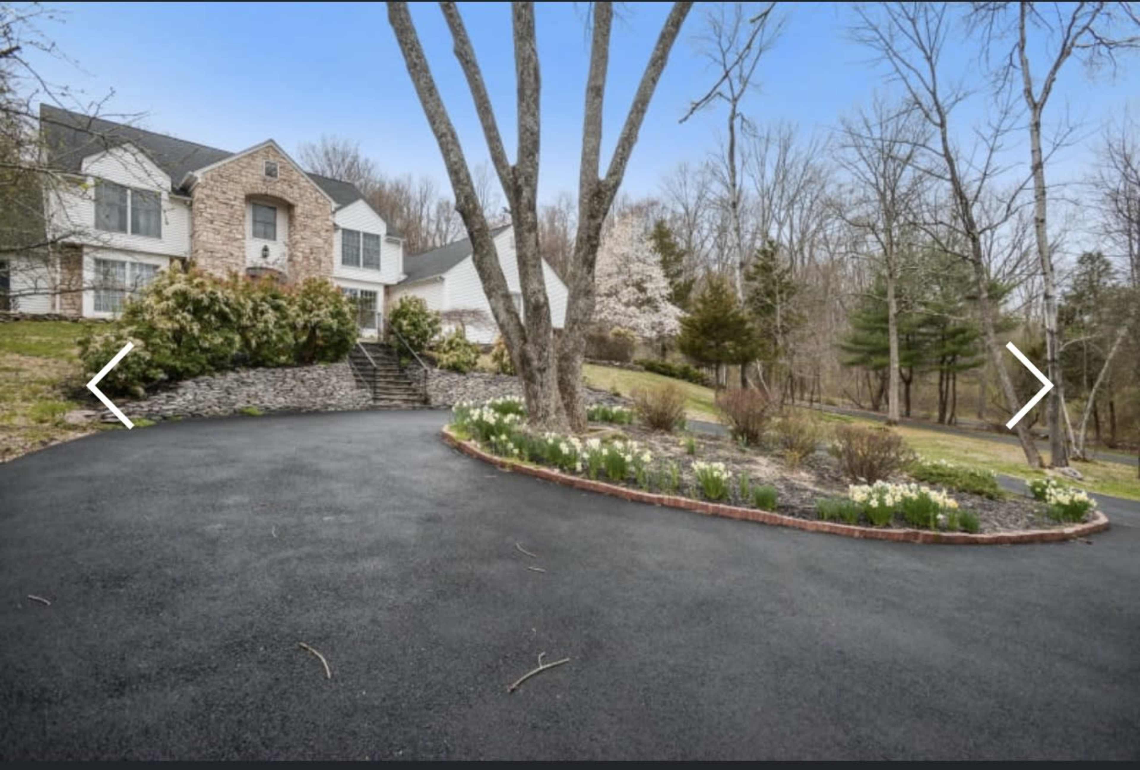 A circular driveway lined with flowers leads to a large house surrounded by trees and shrubs.