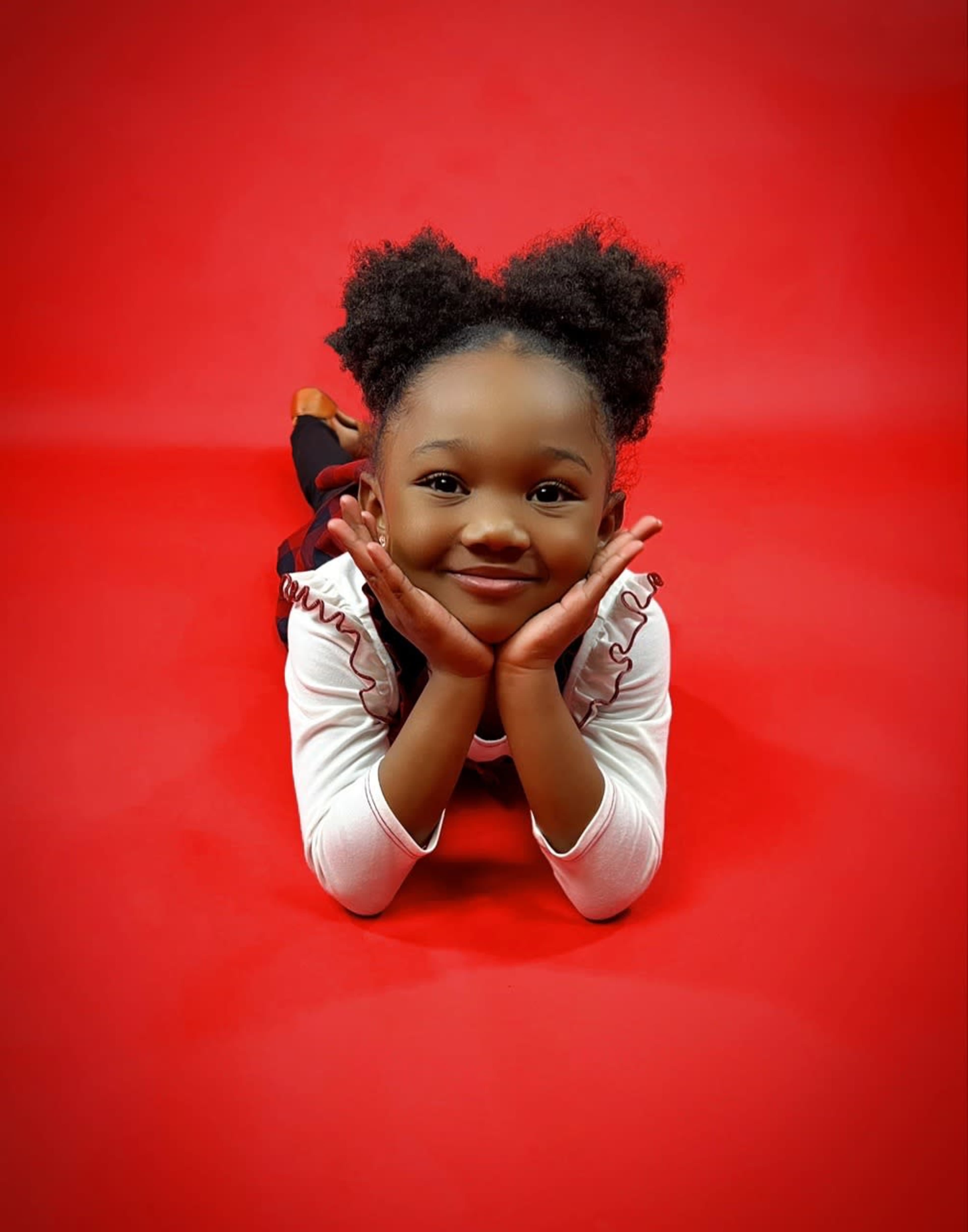 A young girl in a white long-sleeve shirt and black pants lies on a red background, resting her chin on her hands.