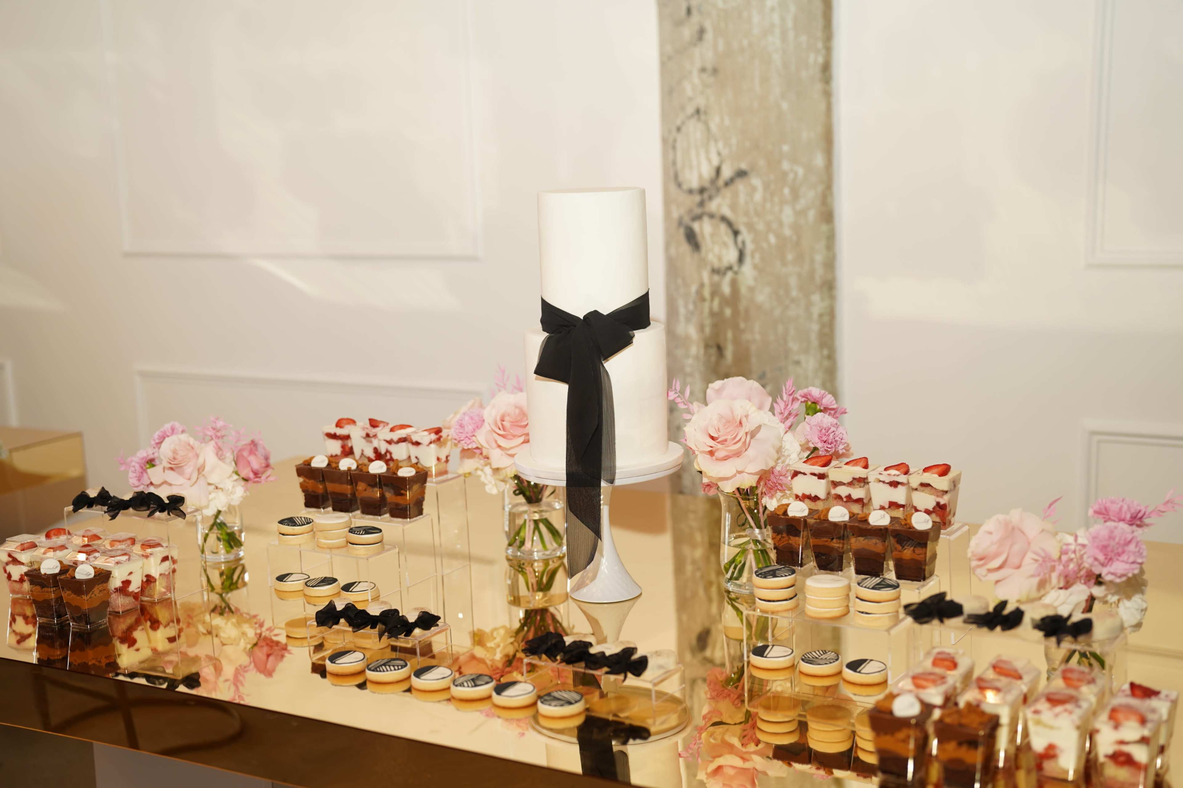 A dessert table features a white cake adorned with a black ribbon, surrounded by delicate floral arrangements and an assortment of mini desserts in glass containers.