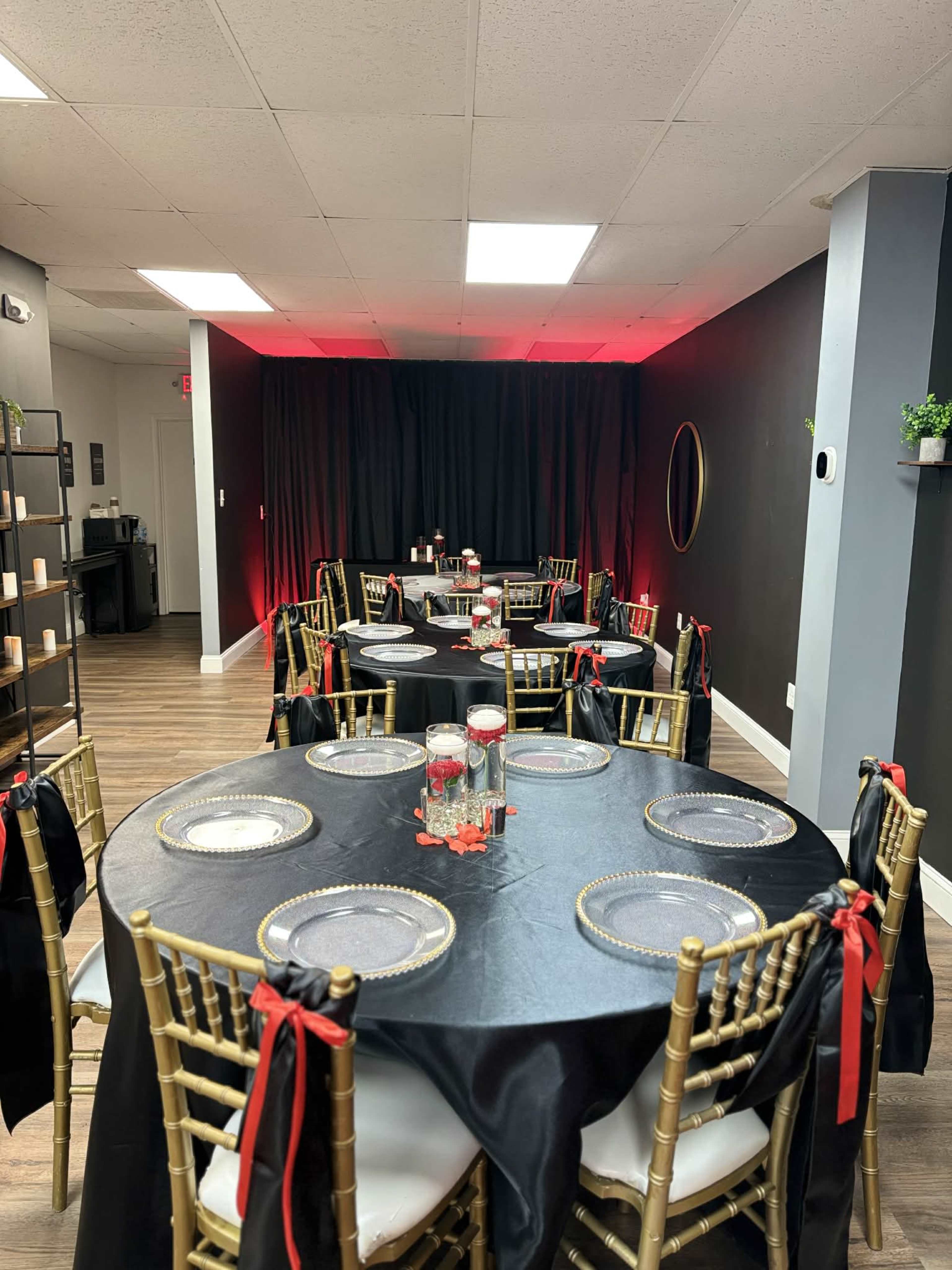 The image shows a dining area with elegantly set tables, featuring black tablecloths, golden chairs, and decorative accents under soft red lighting.
