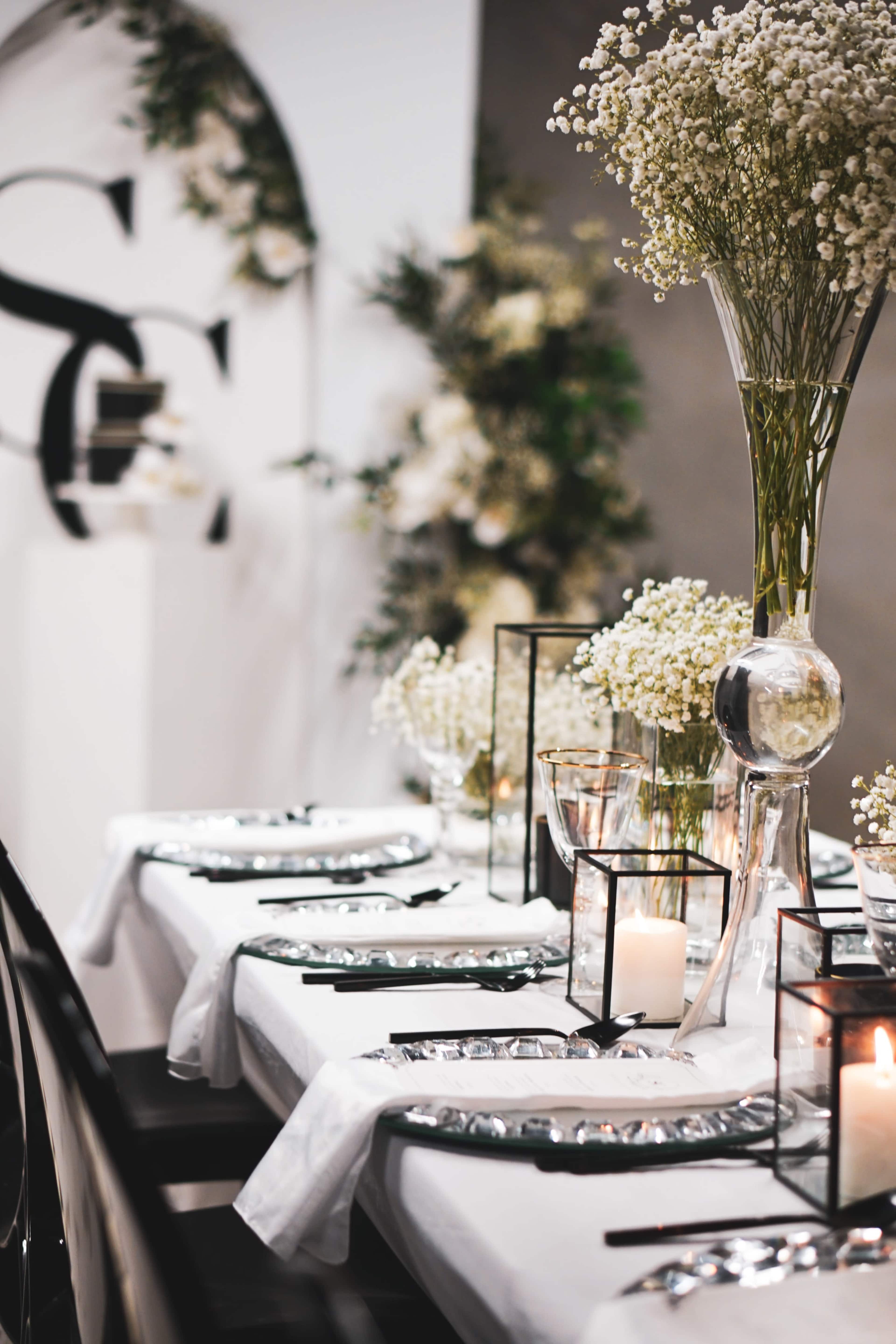 A elegantly set dining table features white tablecloths, glass vases with flowers, candles, and decorative accents in a styled venue.
