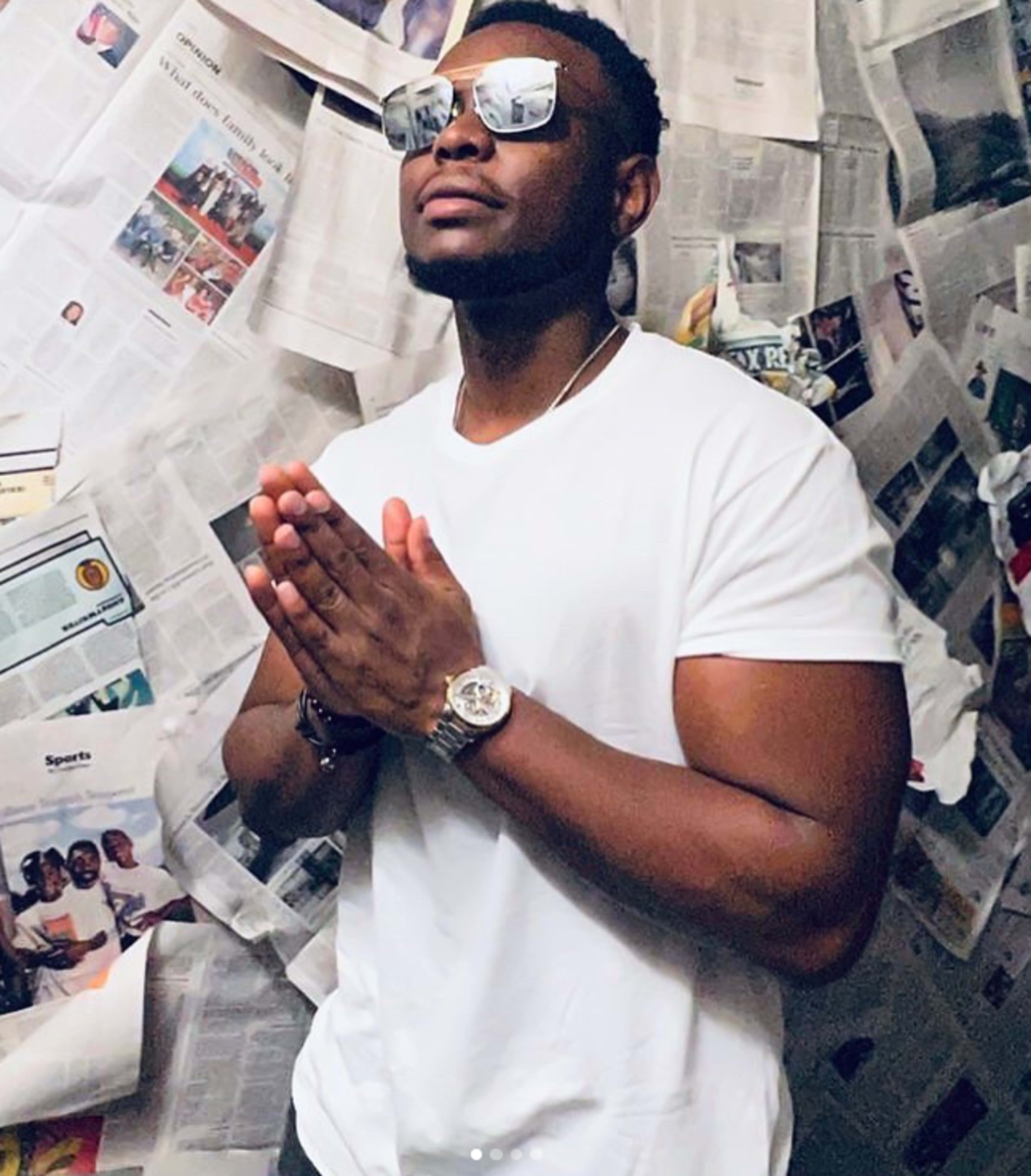 A man in a white t-shirt and sunglasses stands with his hands pressed together in a room covered in newspapers.