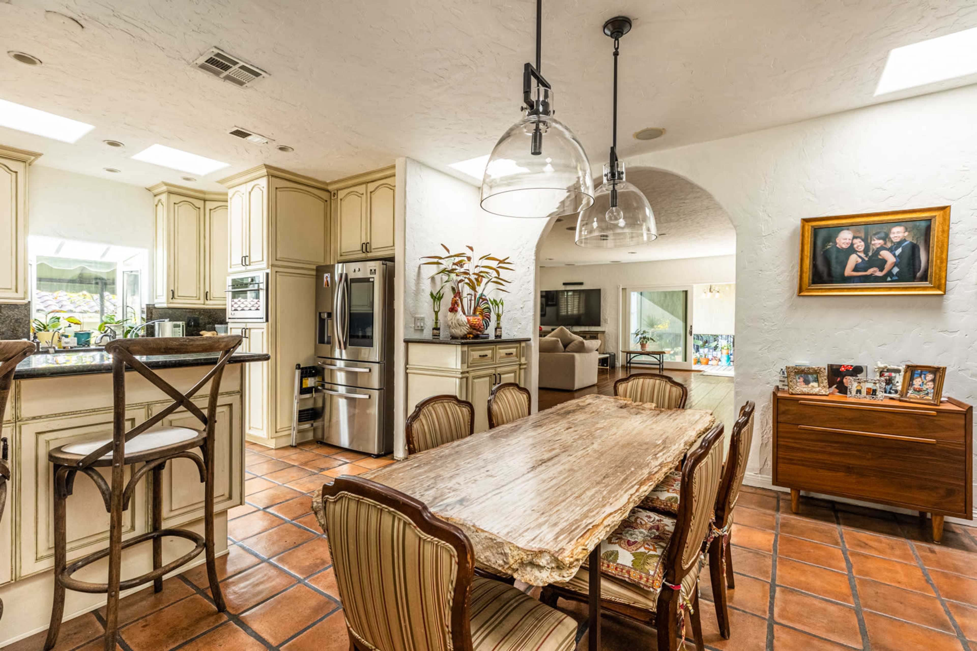 A kitchen and dining area features a wooden dining table surrounded by chairs, with cabinets and a stainless steel refrigerator in the background.