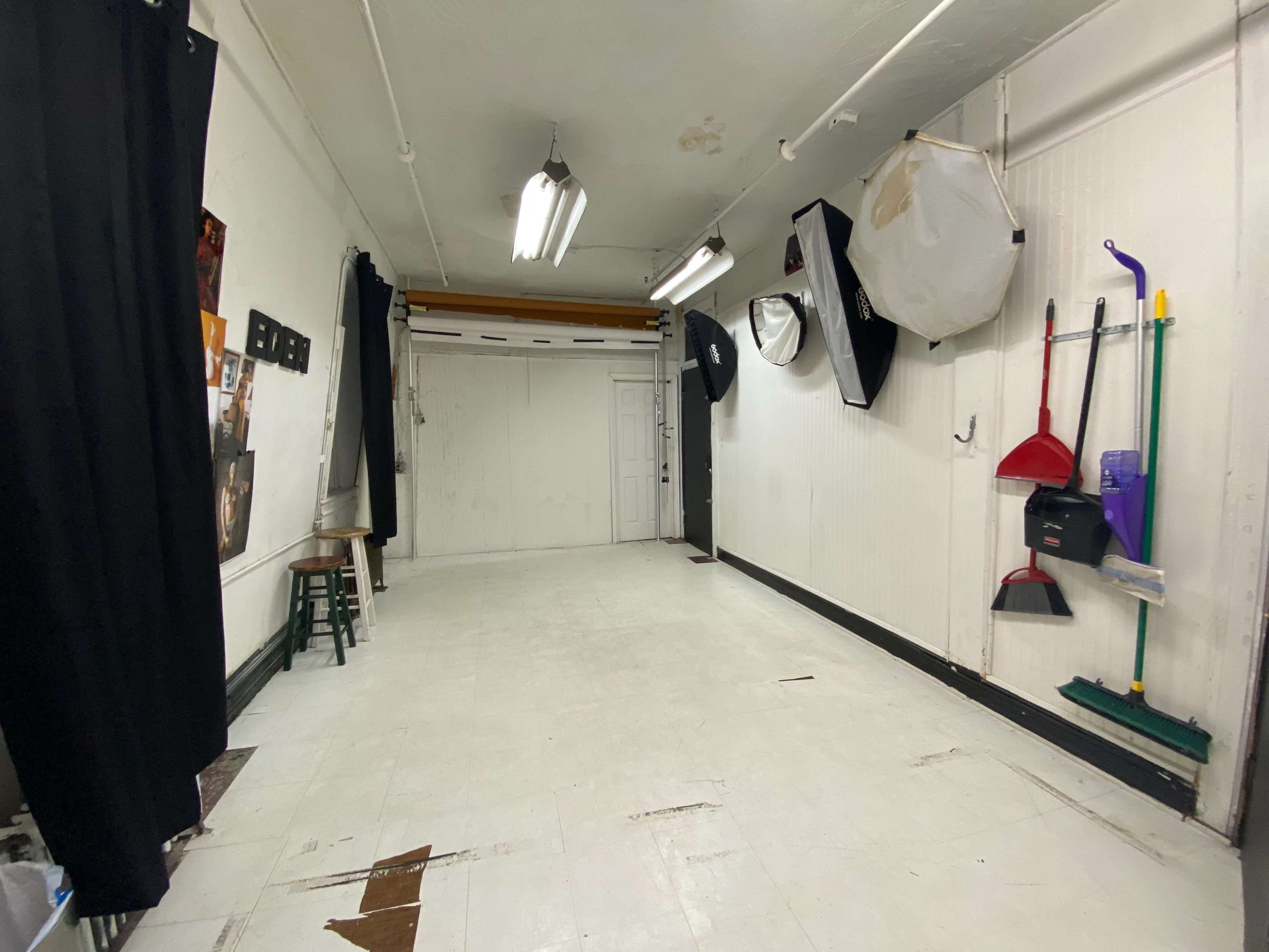 A spacious, empty room with white walls, a wooden stool, a closed door, and several cleaning tools hanging on the wall.