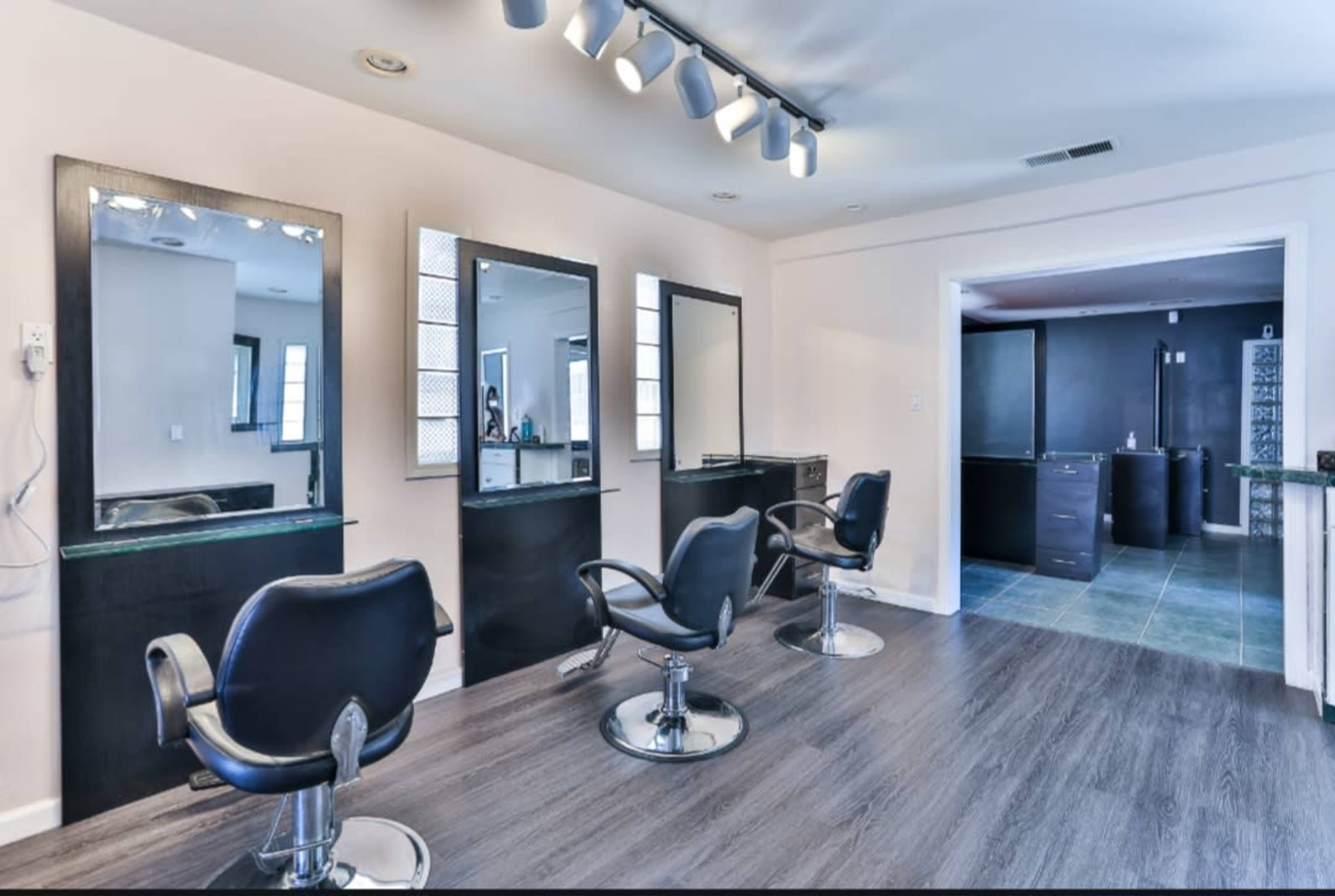A modern hair salon featuring three styling stations with mirrors and chairs, and a contrasting darker room visible in the background.