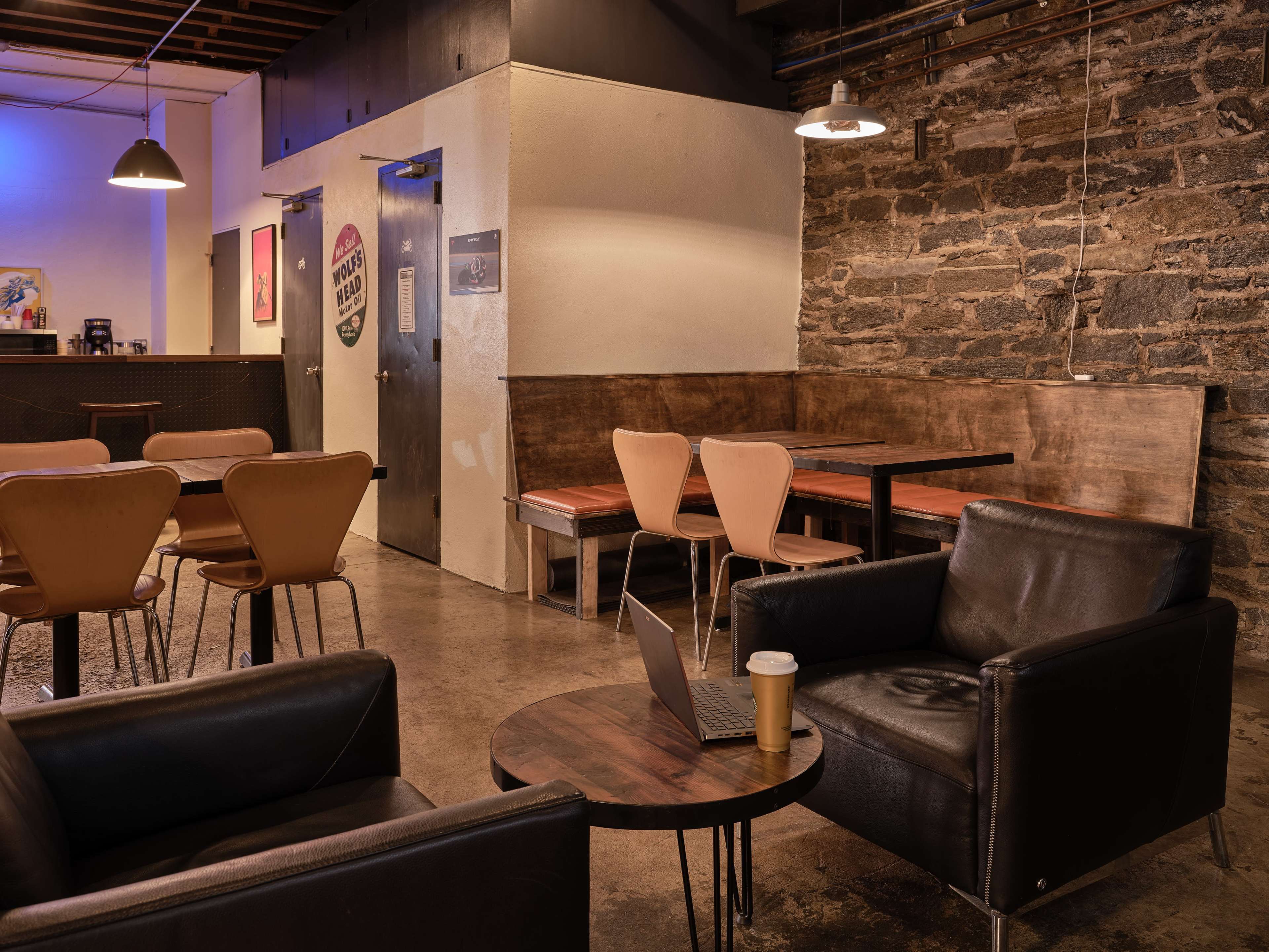 A cozy café interior with leather chairs, wooden tables, and an exposed stone wall, illuminated by overhead lighting.