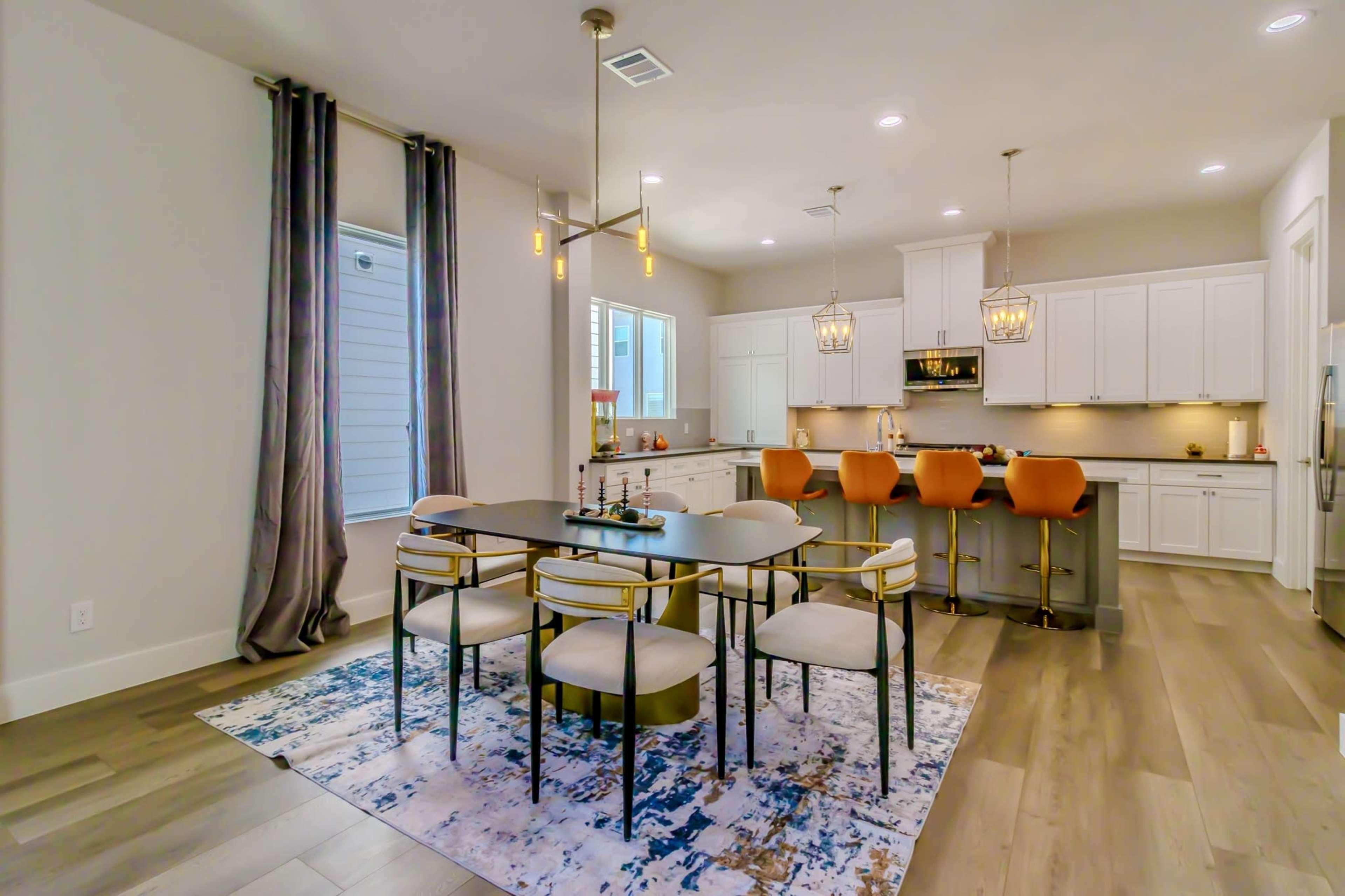 The image shows a modern kitchen and dining area with a large table, six chairs, and bar seating at an island, adorned with a rug and decorative light fixtures.