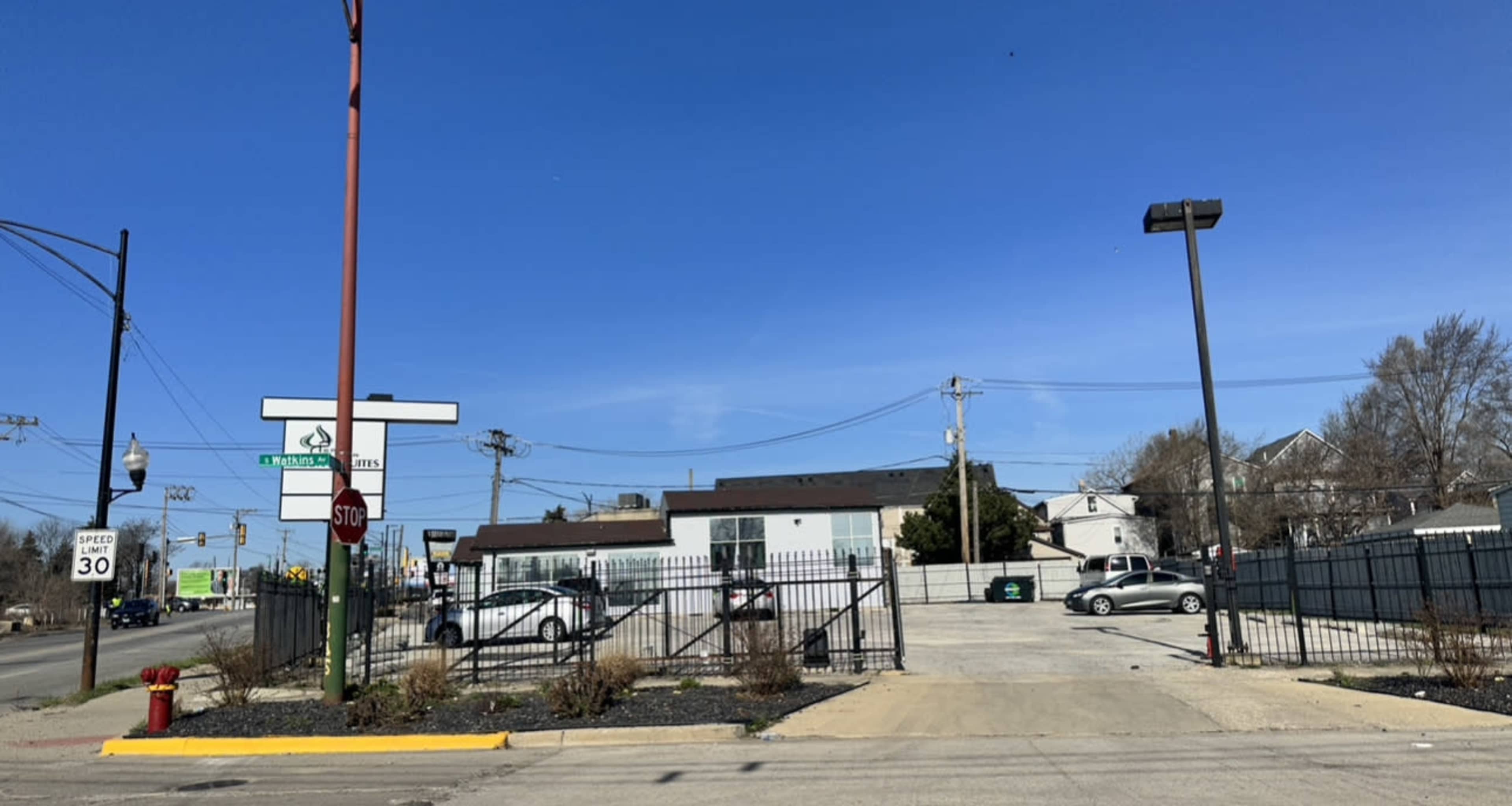 A parking lot adjacent to a street, with a stop sign and various buildings in the background under a clear blue sky.