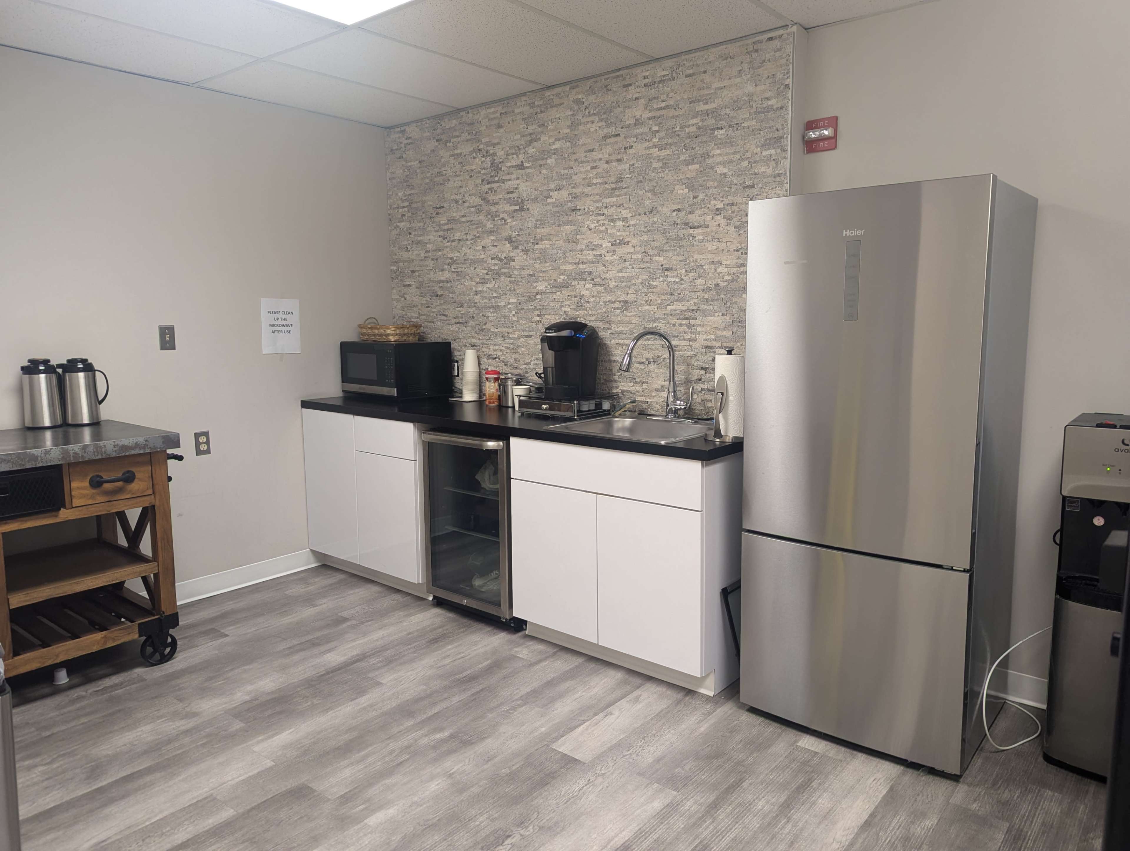 The image shows a modern kitchen space featuring a stainless steel refrigerator, a sink, a coffee maker, a microwave, and a glass-front cabinet, all set against a stone accent wall.