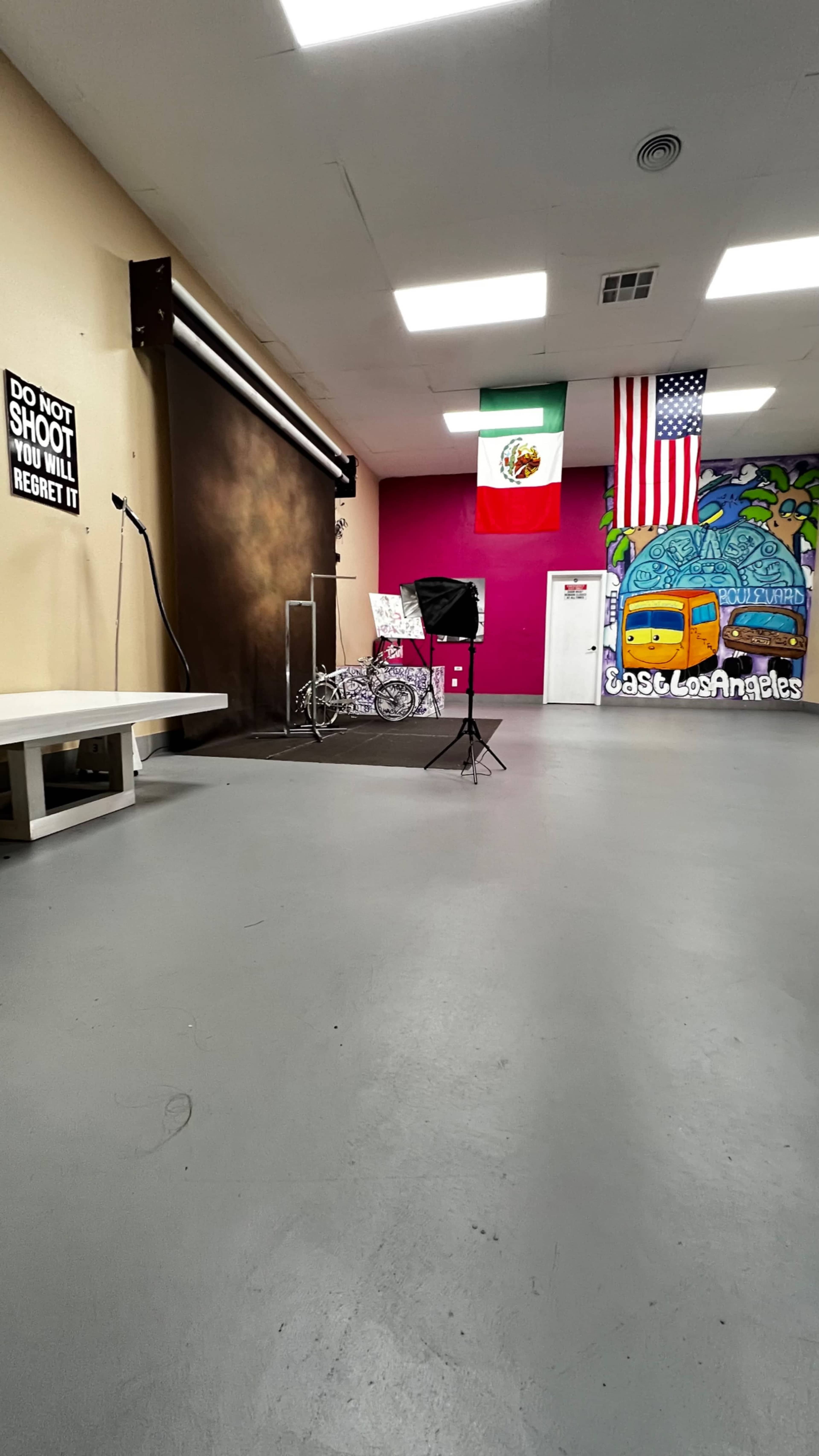 The image shows a studio space with a seamless backdrop, props including a bicycle, a photography light stand, and colorful murals on the walls featuring a Mexican flag and an American flag.