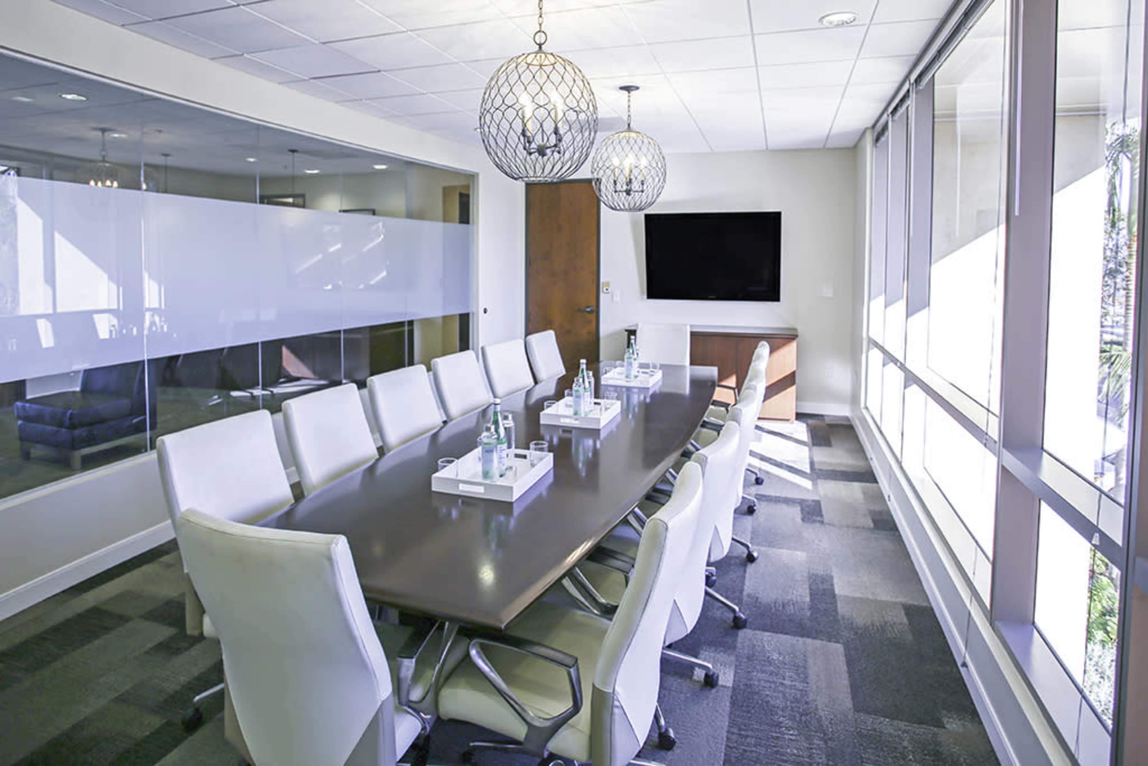 The image shows a modern conference room with a long table, white chairs, and large windows letting in natural light.