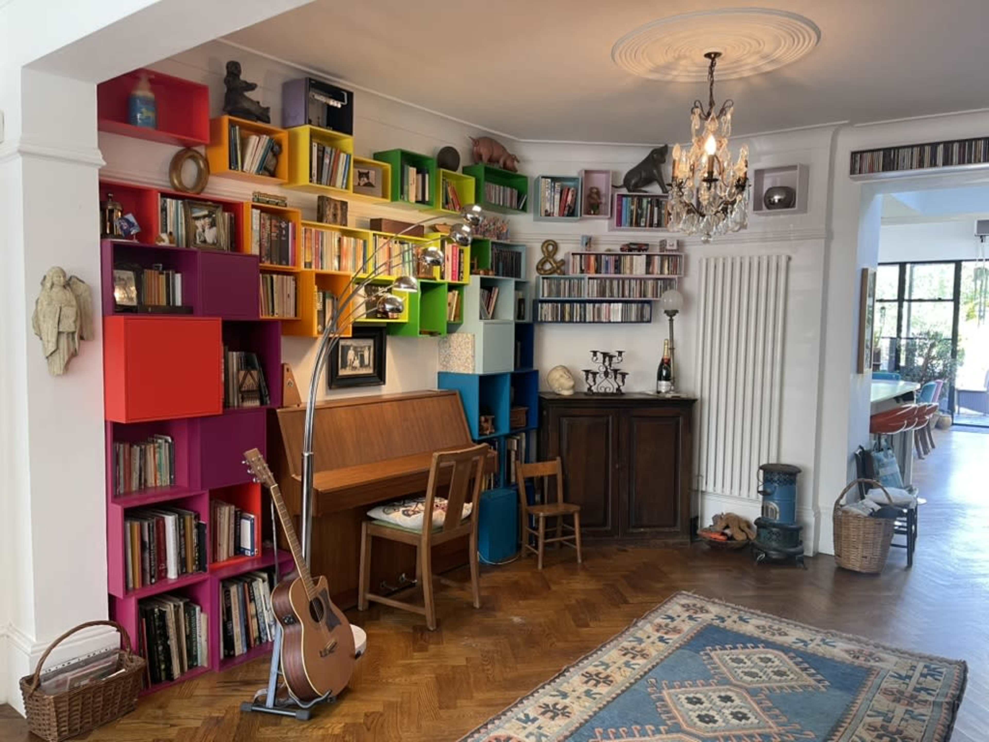The image shows a living room with a colorful bookshelf filled with books, a piano and a small chair, a chandelier overhead, and a cozy rug on the wooden floor.