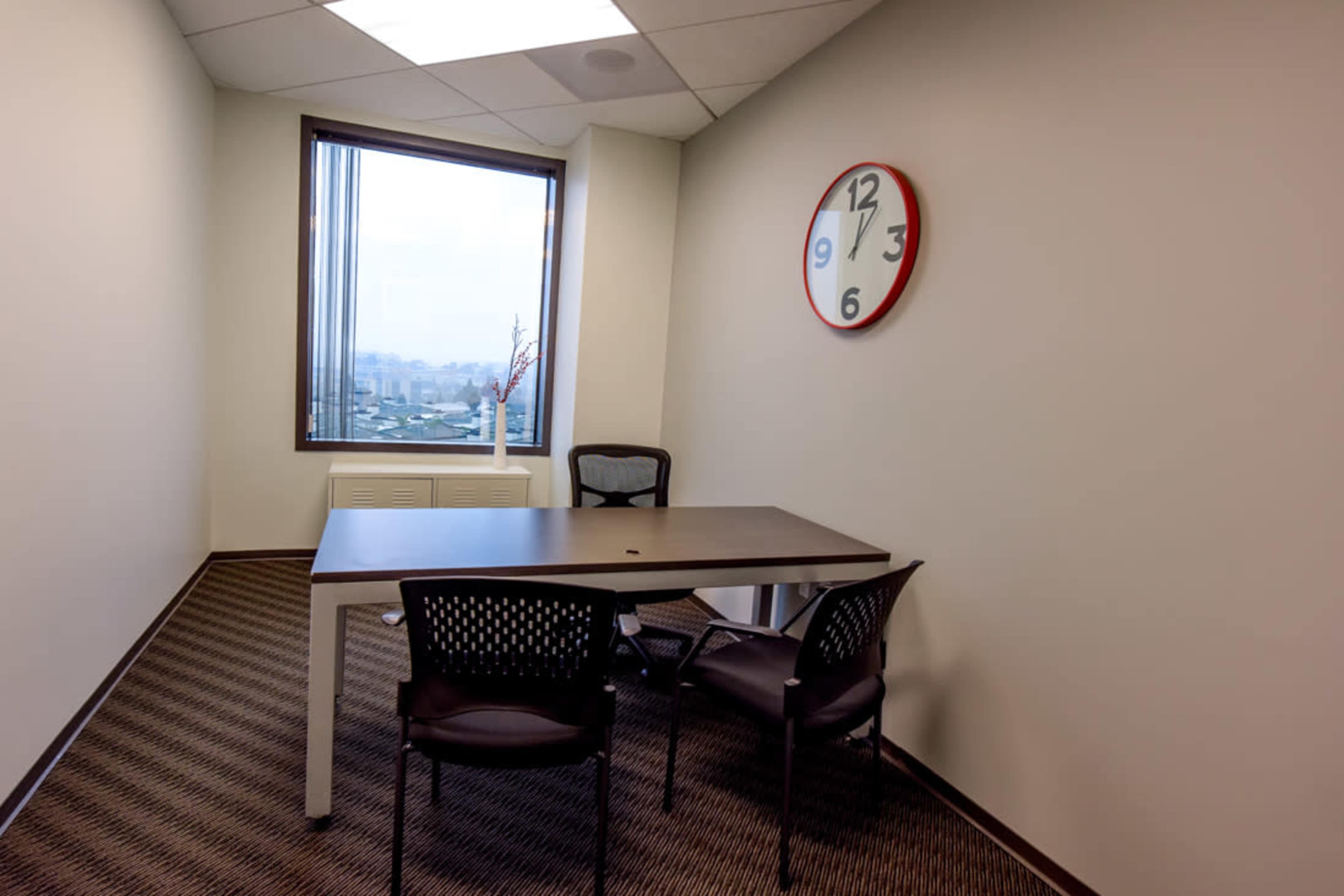 A small office meeting room featuring a table, two chairs, a wall clock, and a window with a view.