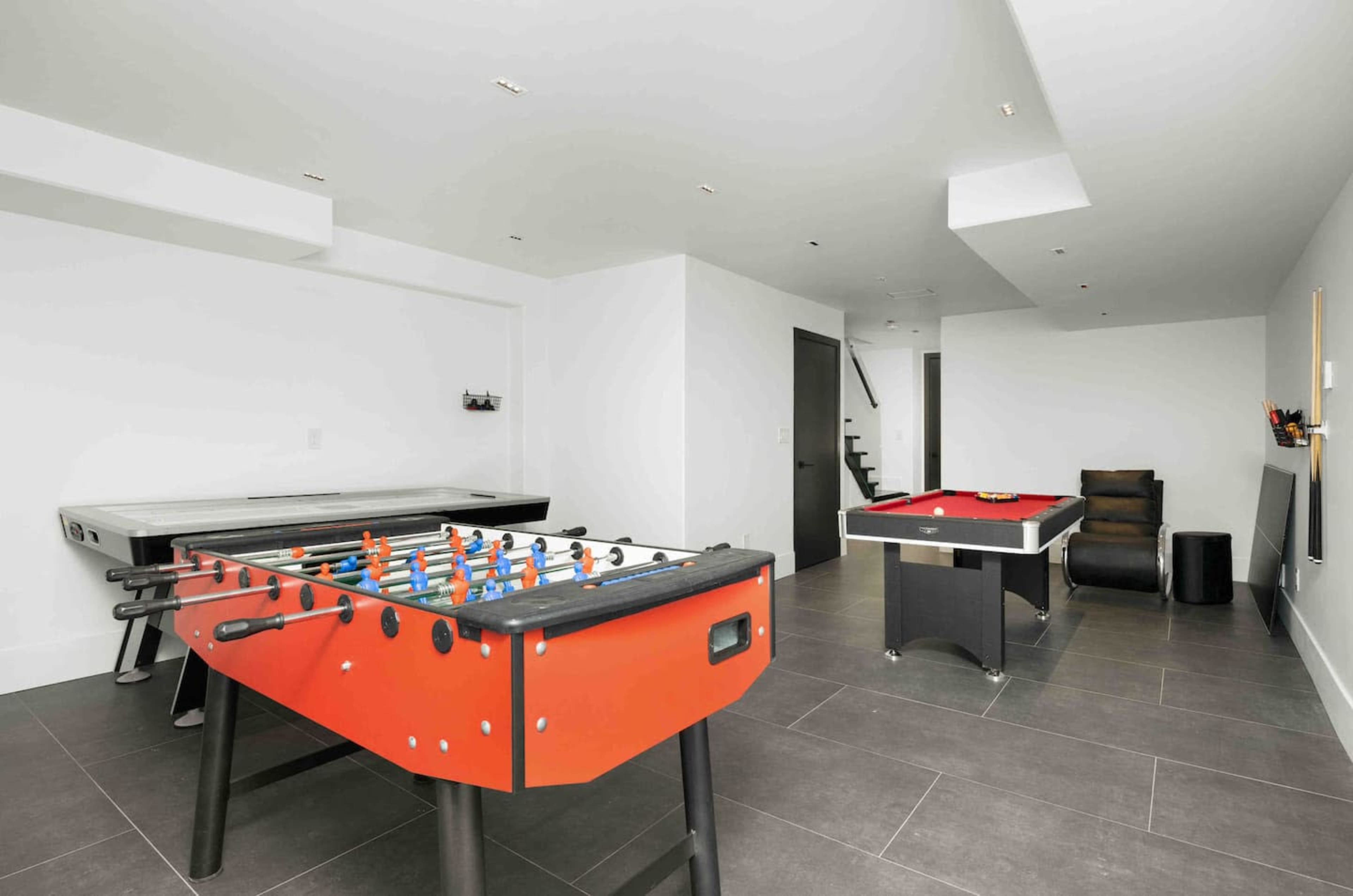 The image shows a game room containing a foosball table, a pool table, and a black chair, all set on tiled floors.