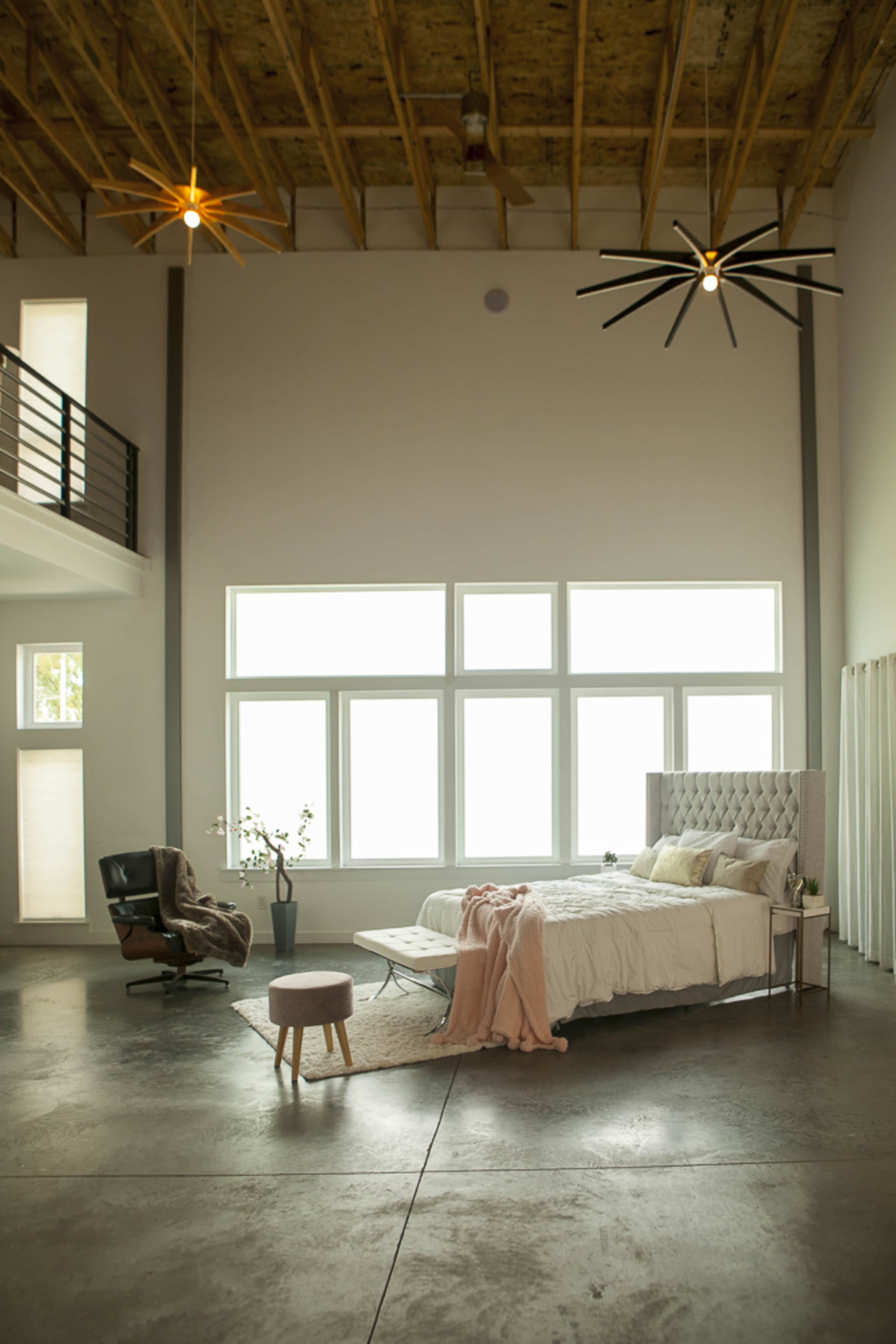 A modern bedroom features a large bed with a tufted headboard, a comfortable chair, and large windows that allow natural light to flood the space.