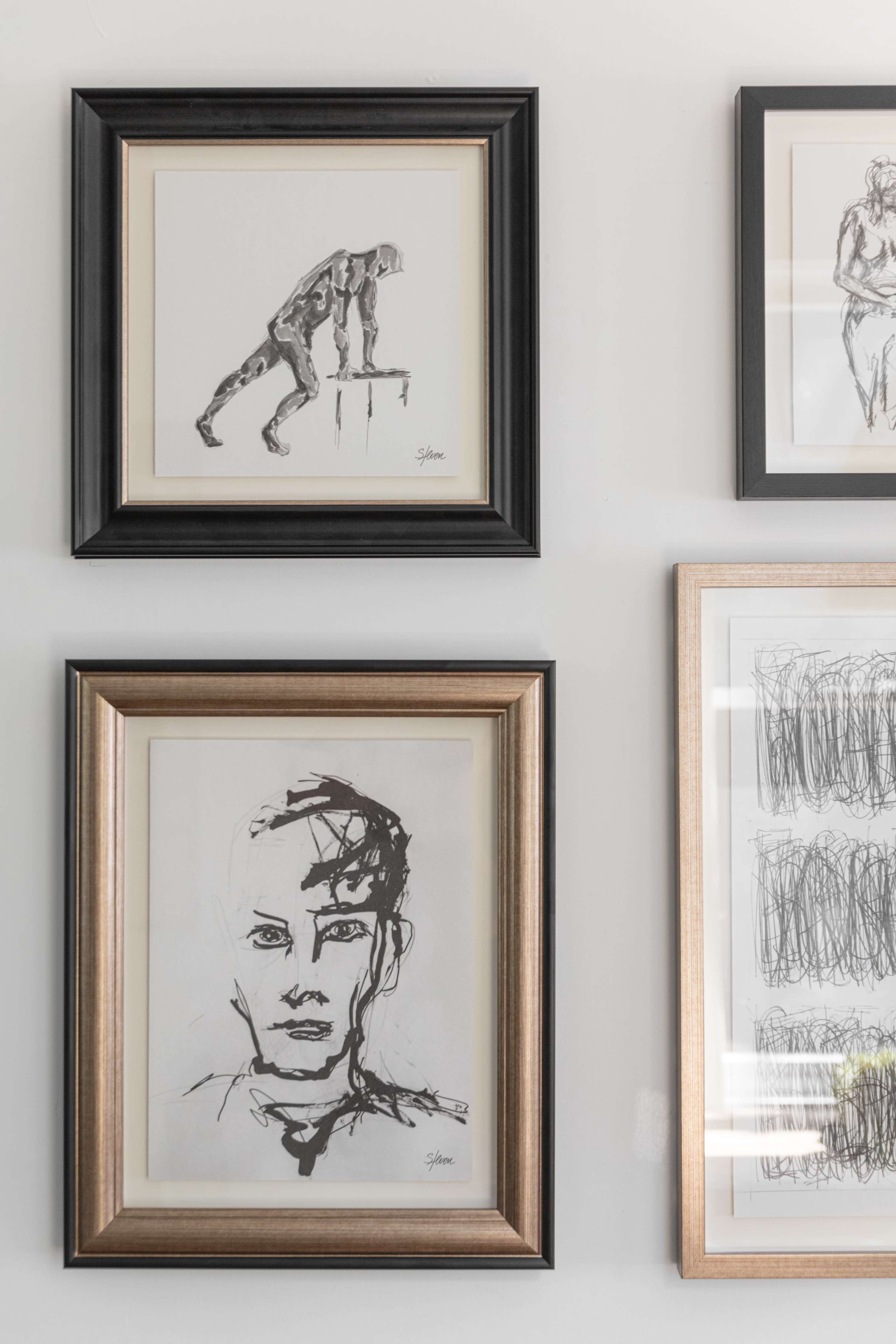 A collection of framed sketches, including a figure leaning on a surface and a portrait of a person, displayed on a wall.
