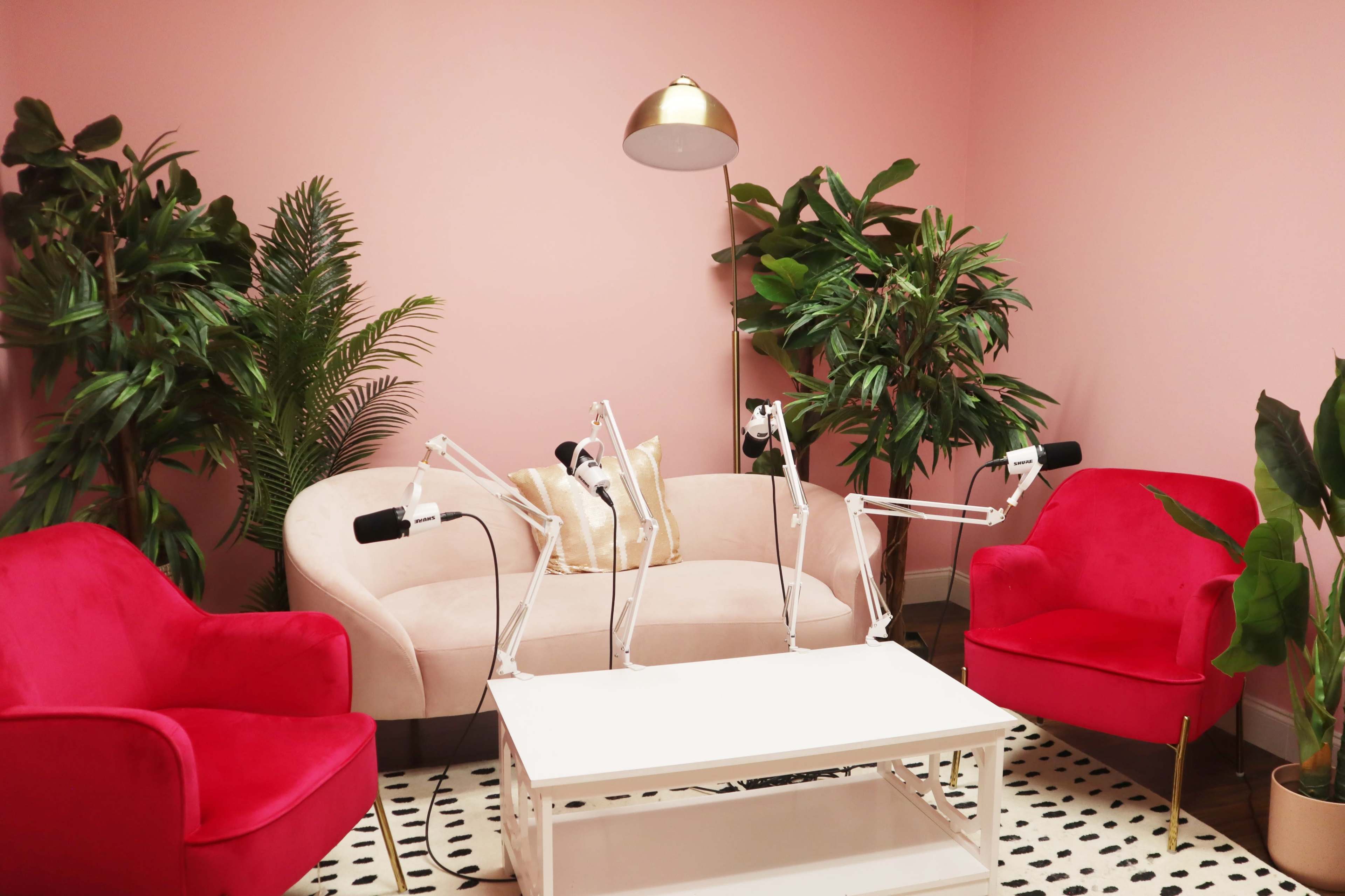 A modern seating area features a light pink sofa and two bright red armchairs, surrounded by greenery and a white coffee table with microphones for recording.