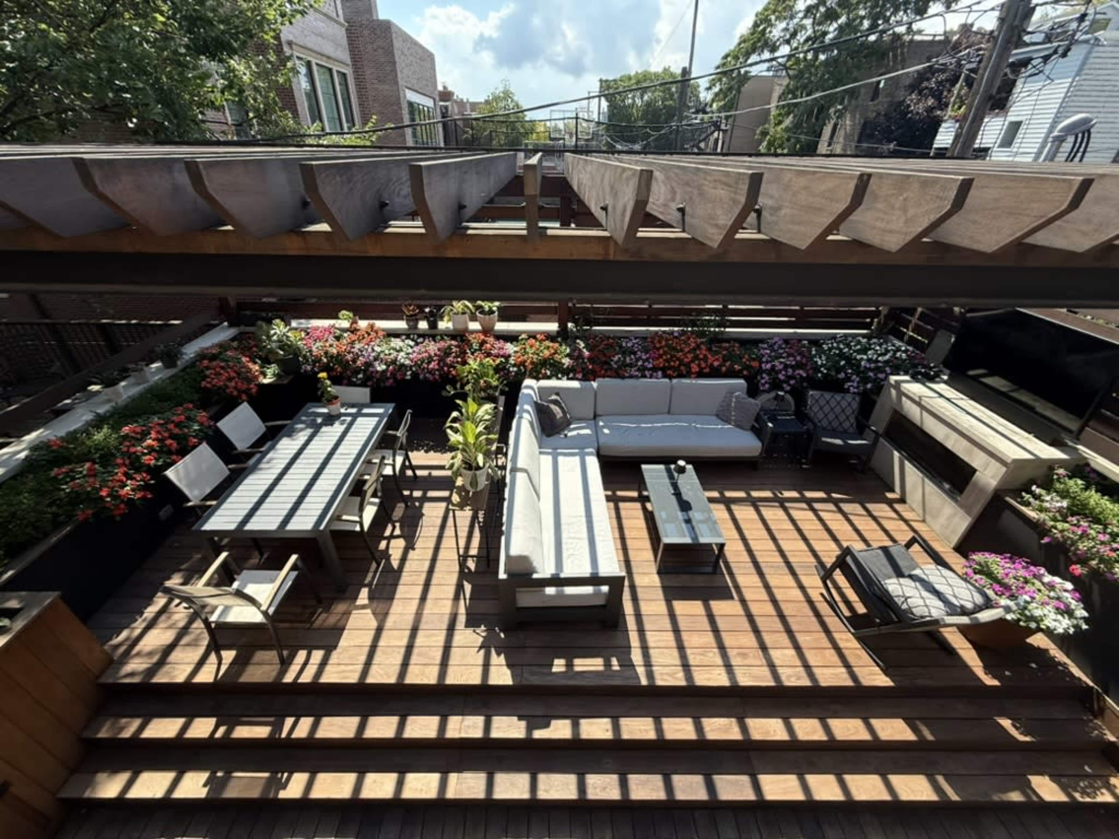 The image shows a spacious rooftop patio adorned with various colorful flowers, featuring seating areas, a dining table, and a wooden deck.