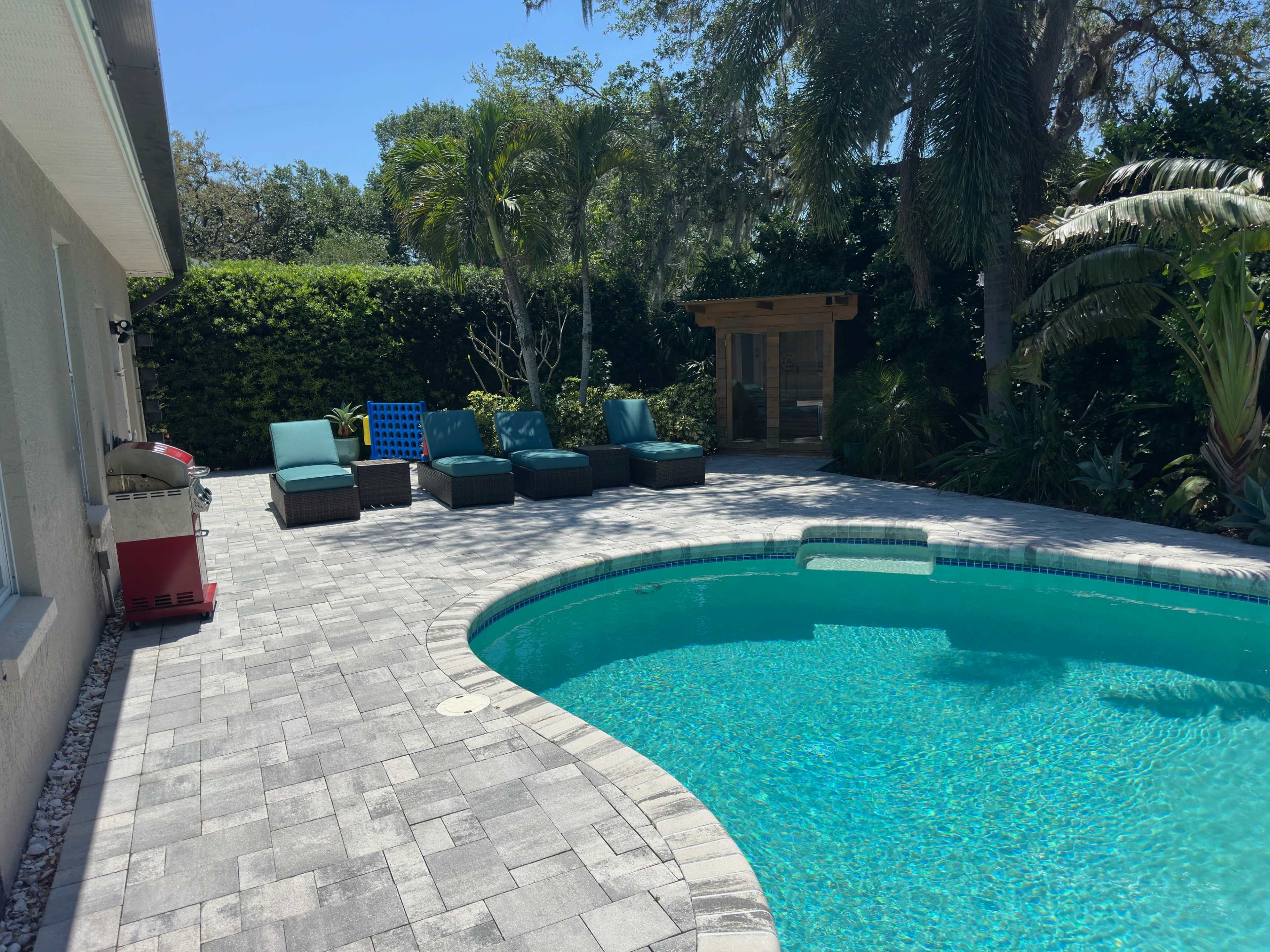 A clear swimming pool is surrounded by a patio featuring lounge chairs and a nearby shaded area with landscaping.