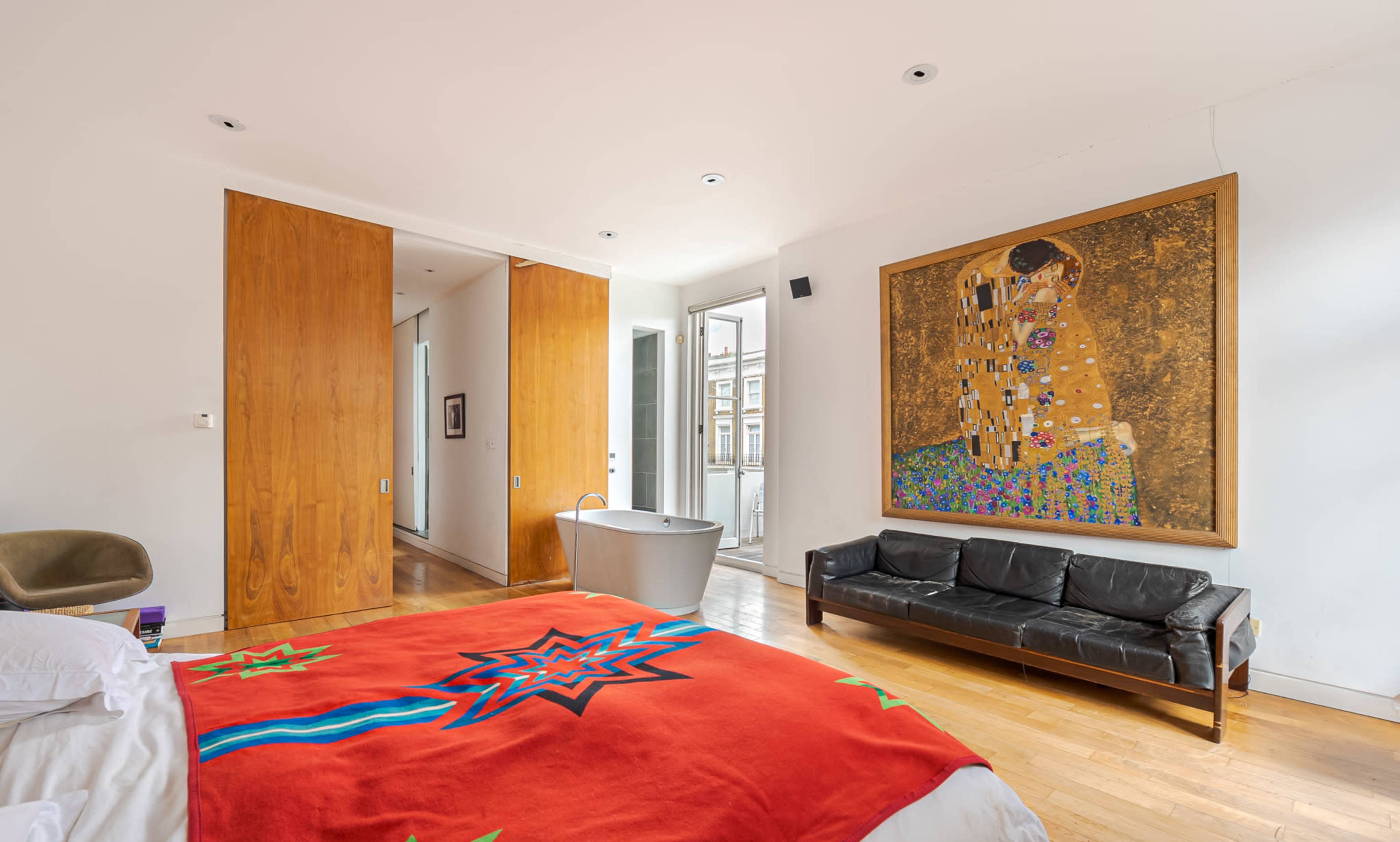 A modern bedroom features a large bed with a red and blue coverlet, a stylish black couch, a freestanding bathtub, and a prominent painting on the wall.